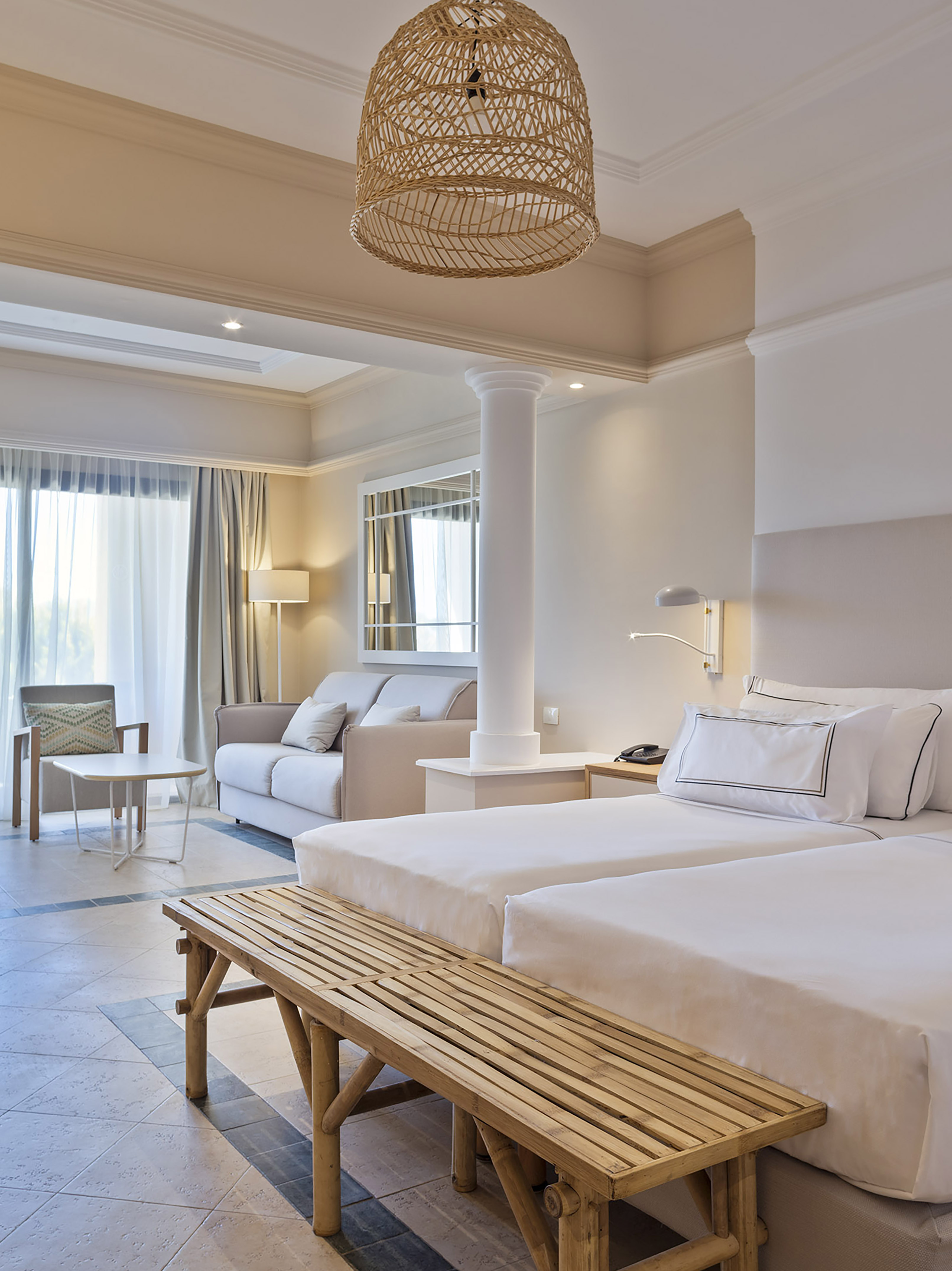 Bright hotel suite with twin beds, bamboo bench, sofa, and large window.