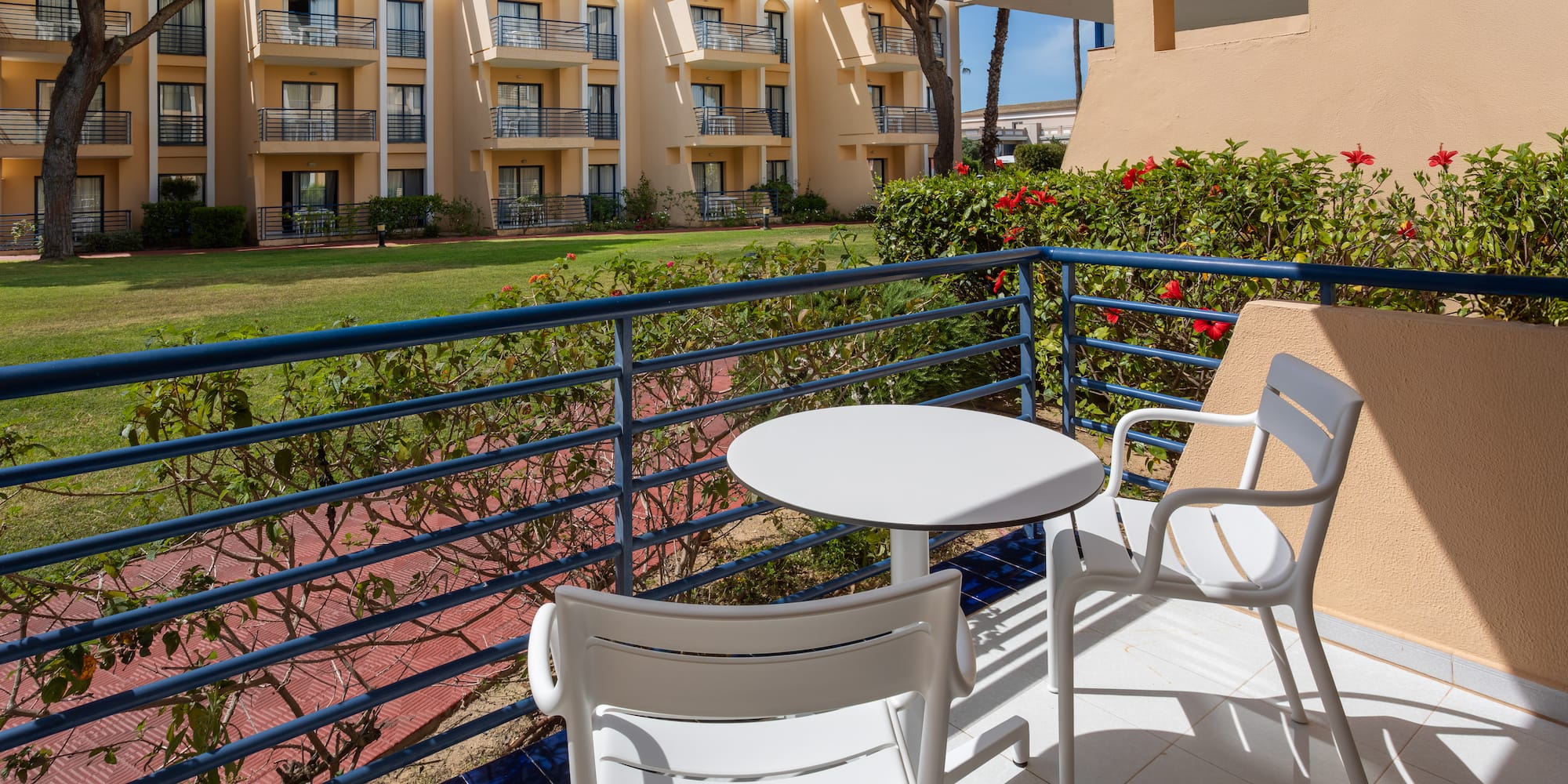Hotel balcony with white chairs, table, garden, and resort building view.