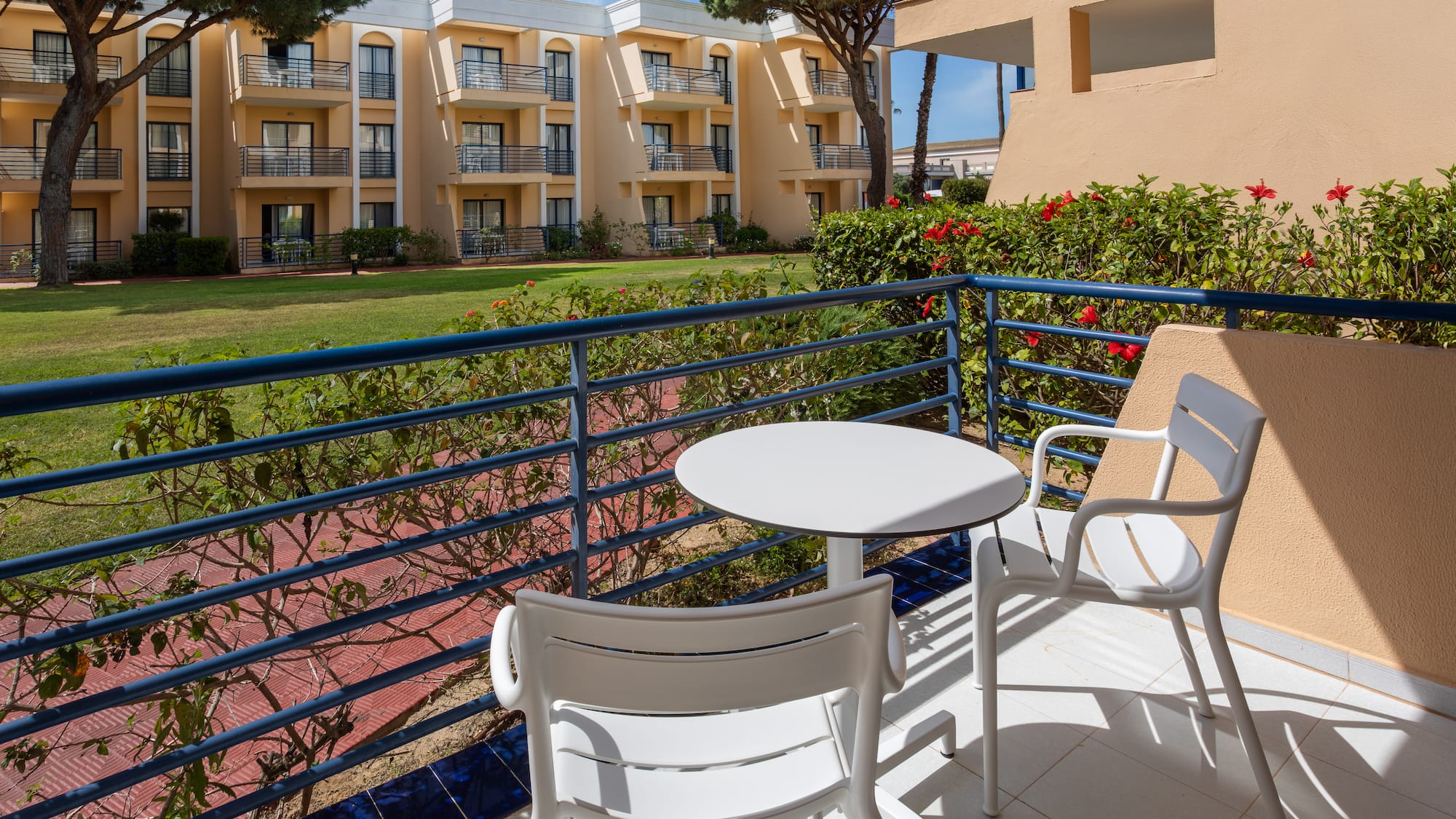 Sunny hotel balcony with white outdoor seating, garden, and resort building.