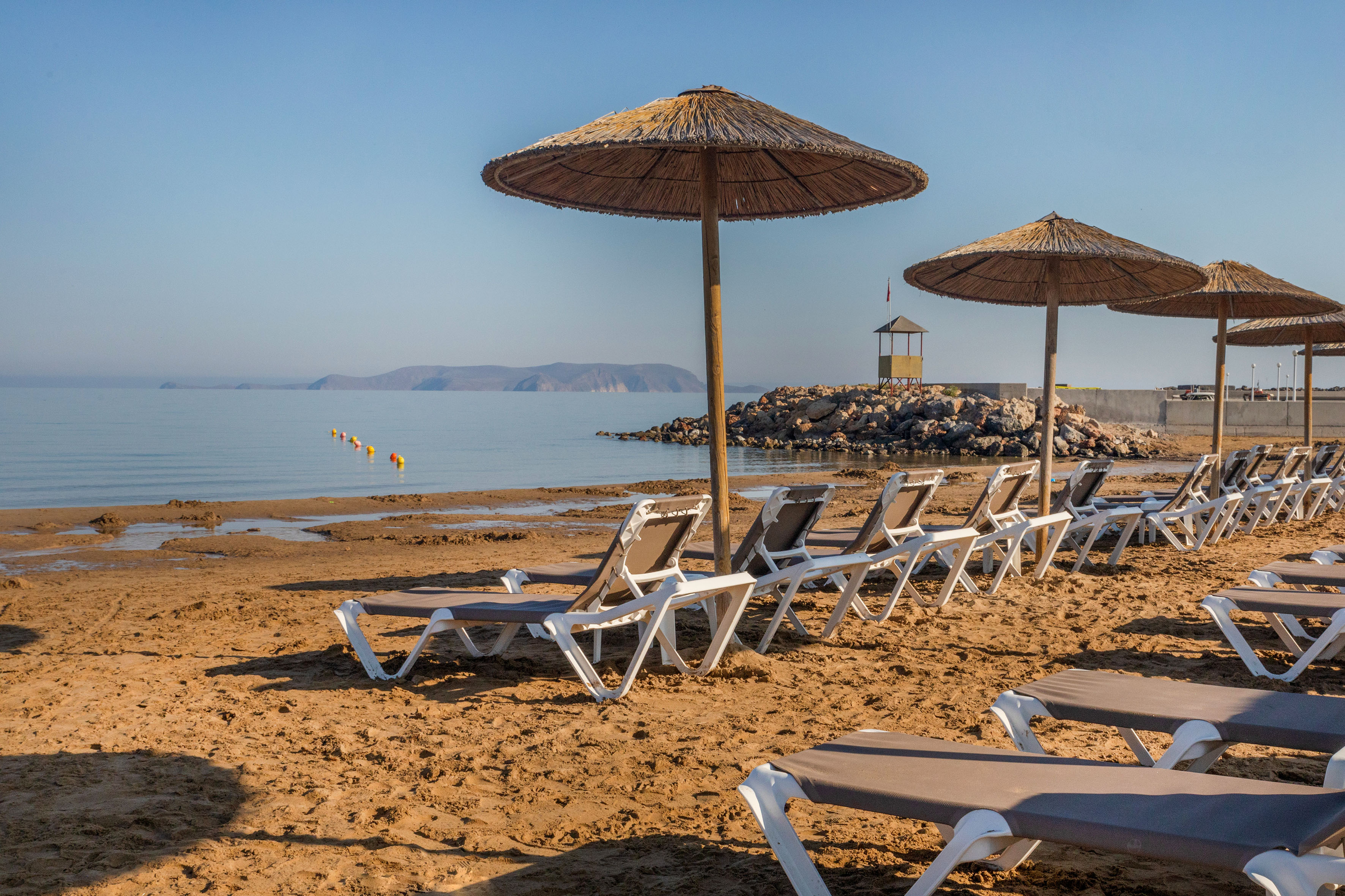Golden sandy beach with sunbeds, straw umbrellas, calm sea, and distant mountains.