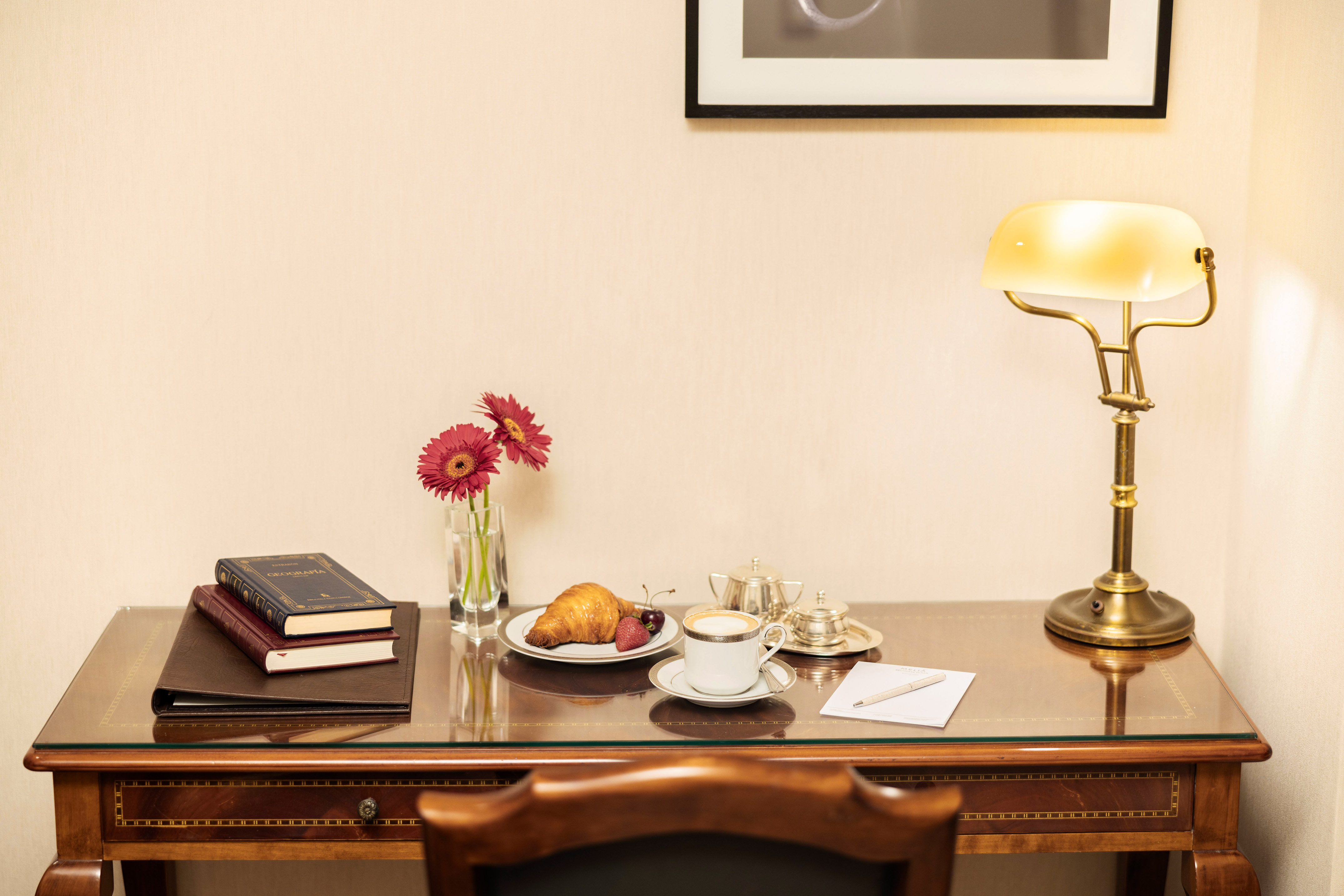 Hotel desk with coffee, croissant, books, red flowers, and a lit brass lamp.