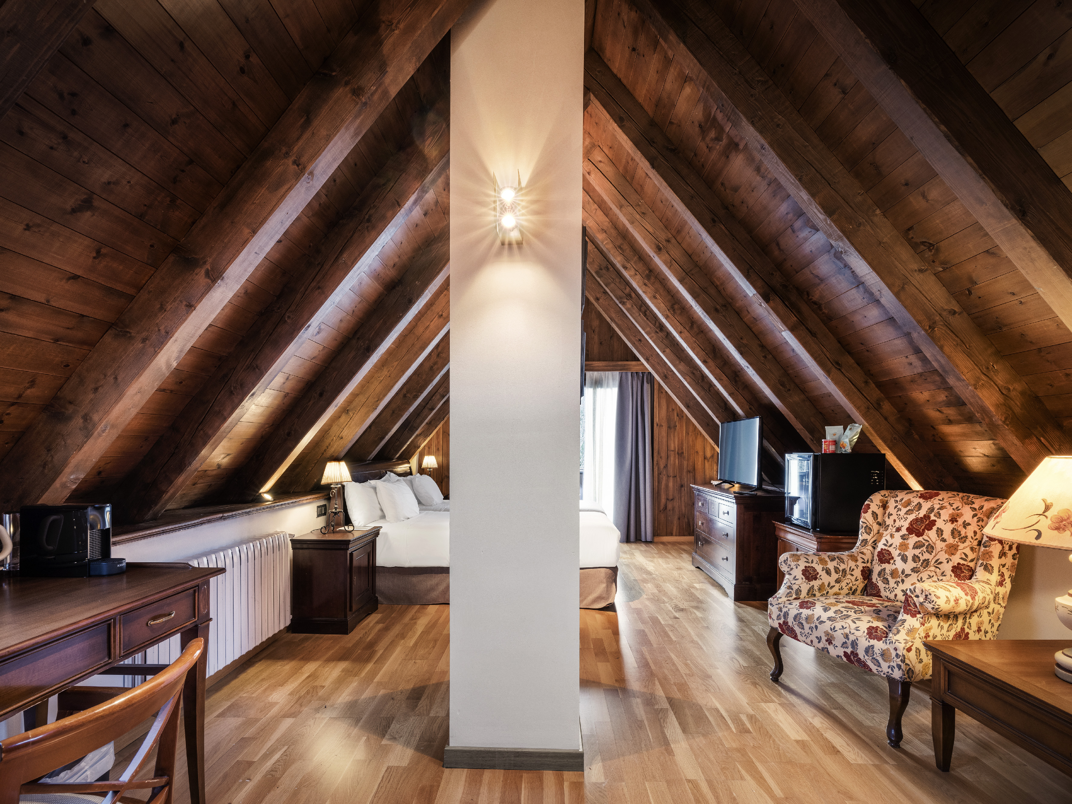 Rustic attic hotel suite with wooden beams, bed, desk, and floral armchair.
