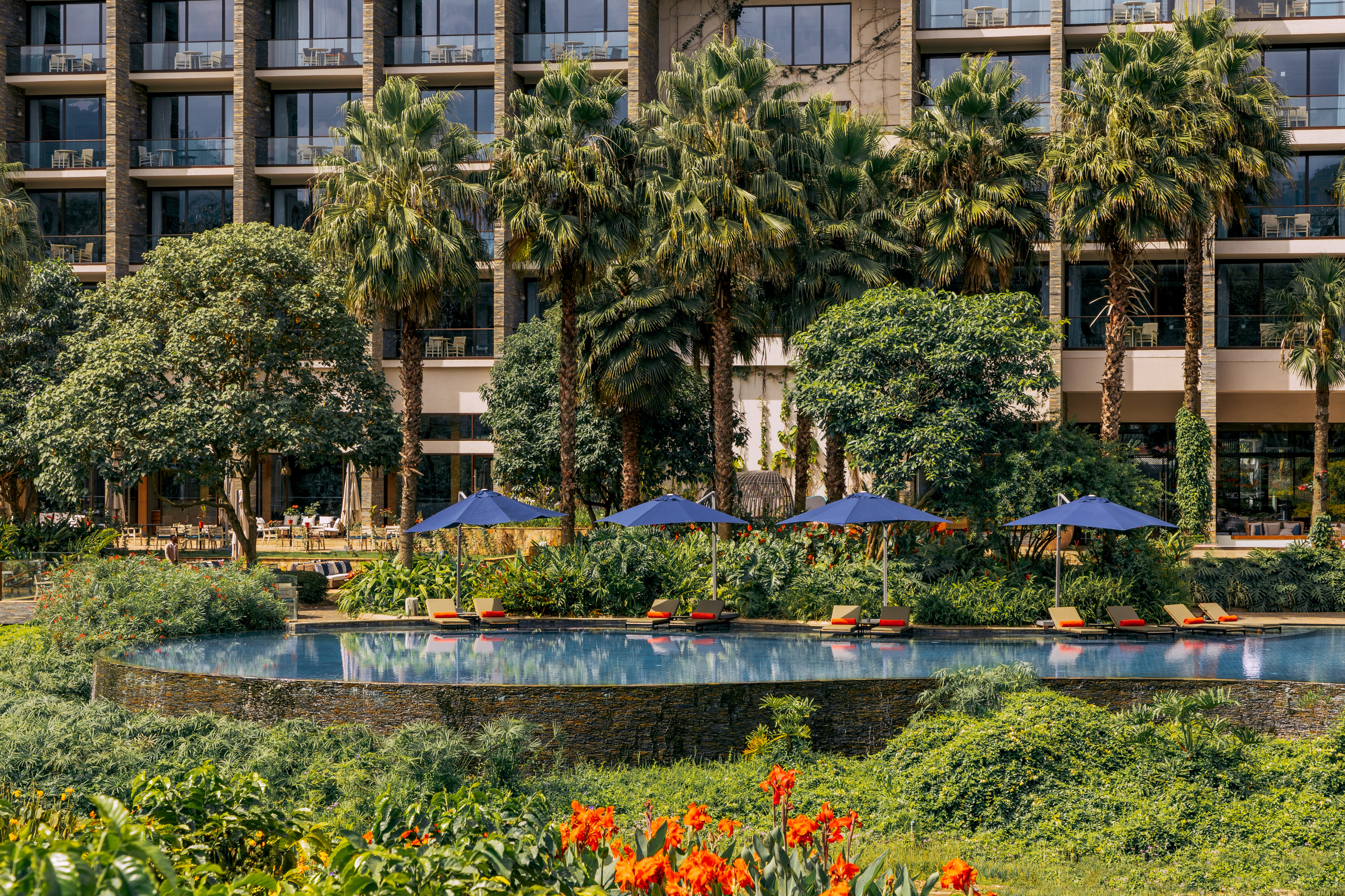 Tropical resort pool with palm trees, blue umbrellas, and orange flowers.