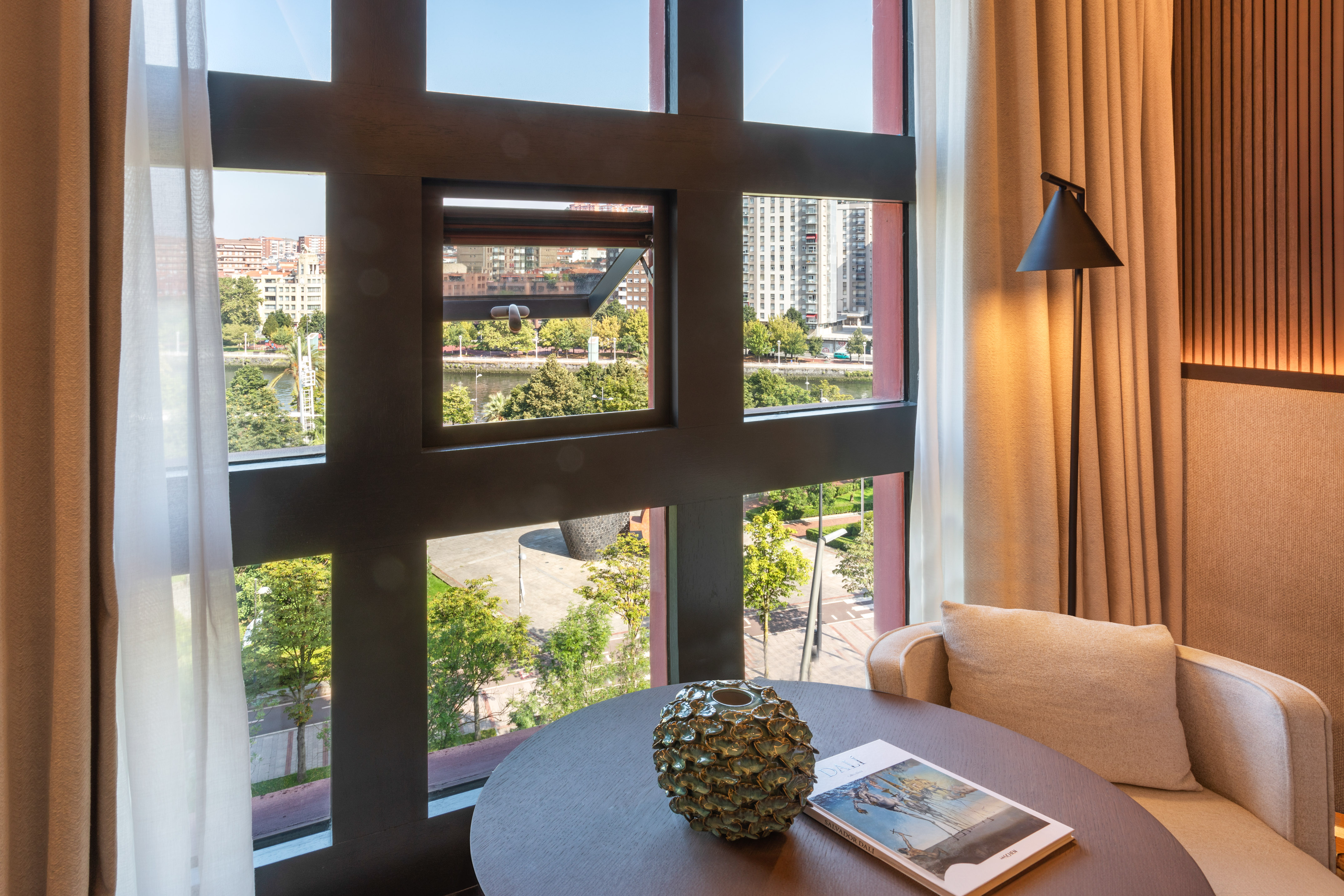 Modern hotel room with large window, city river view, and cozy seating area.