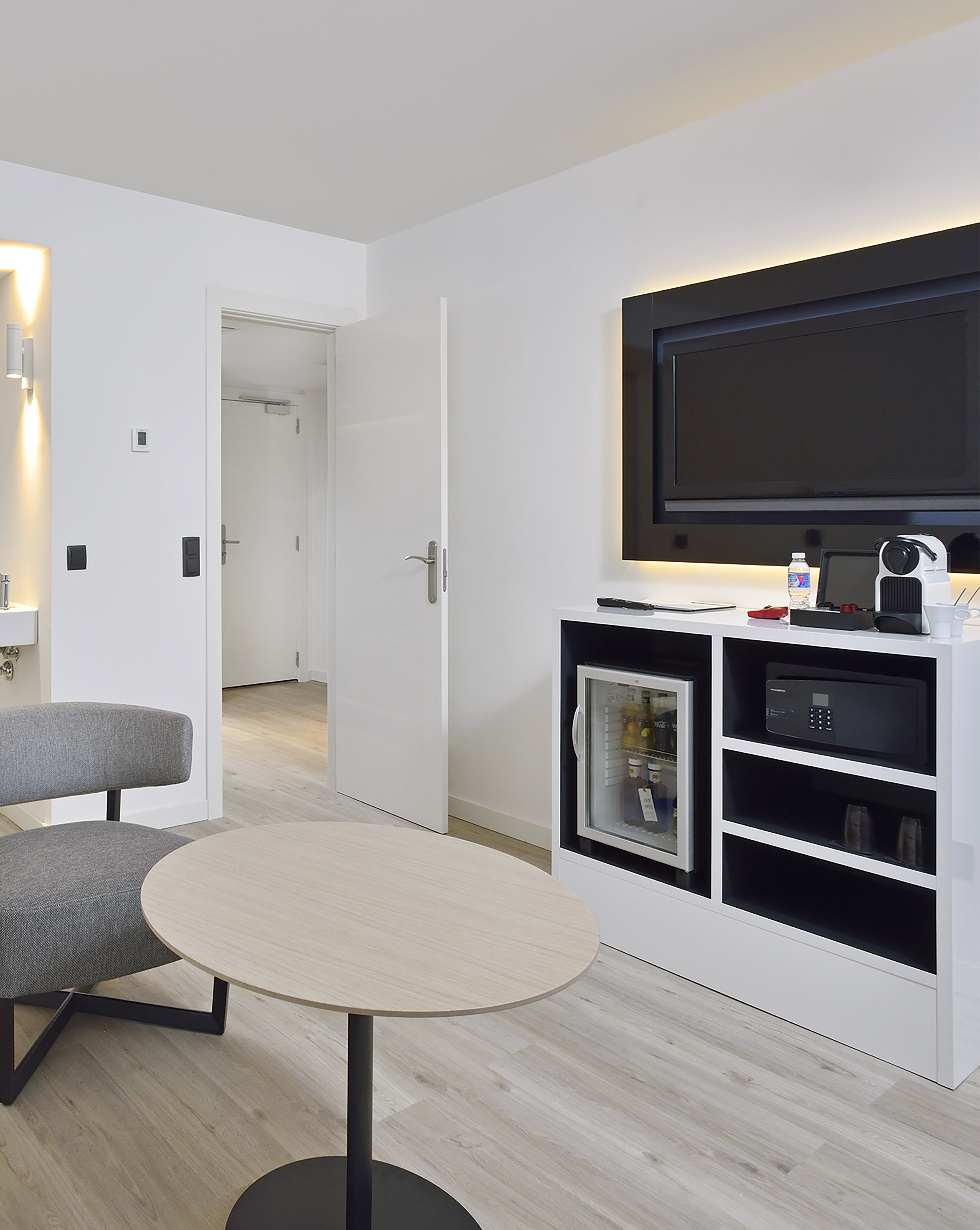 Modern hotel room with TV, mini fridge, safe, coffee machine, and seating.