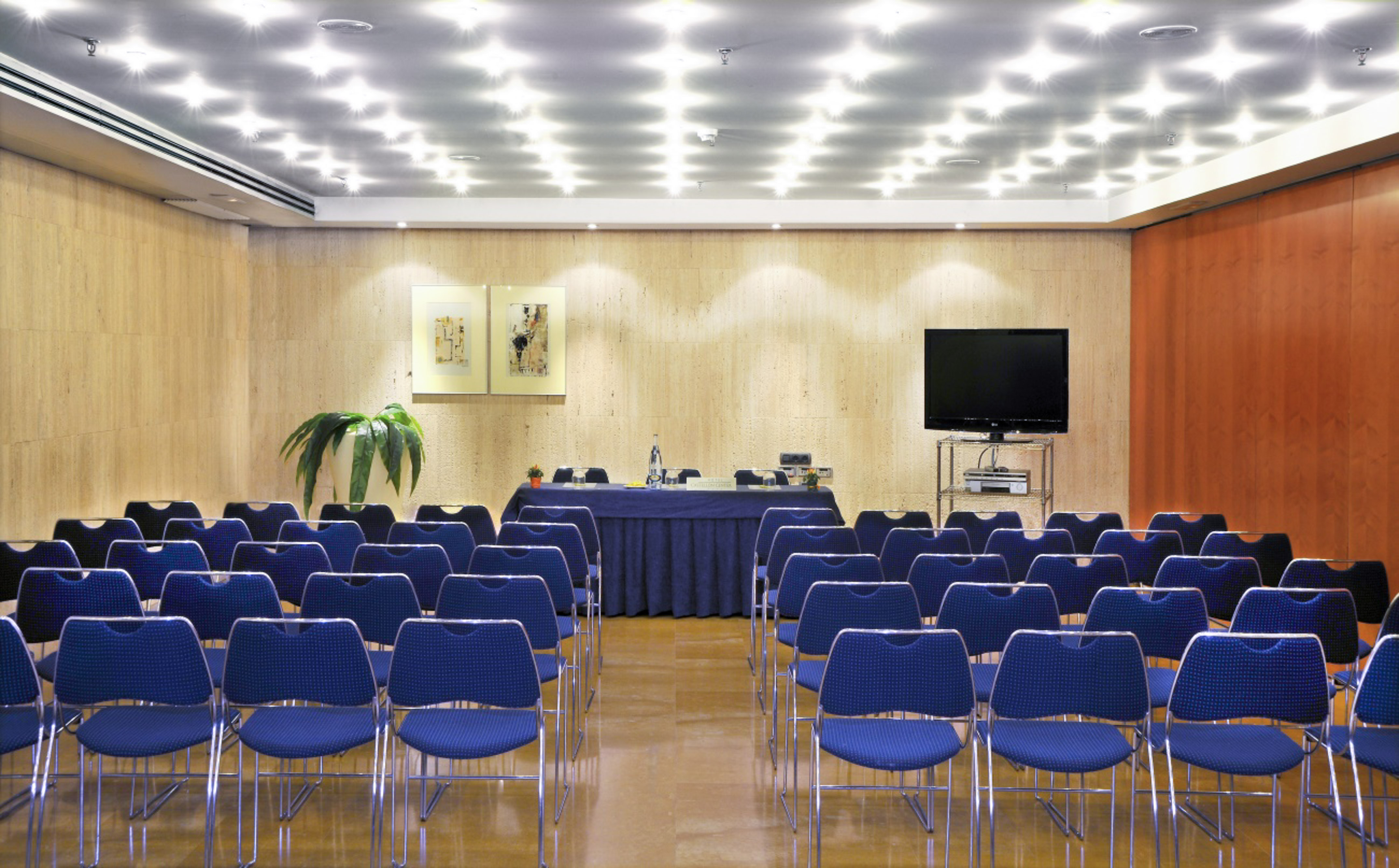 Conference venue with rows of blue chairs, presentation table, and TV screen.