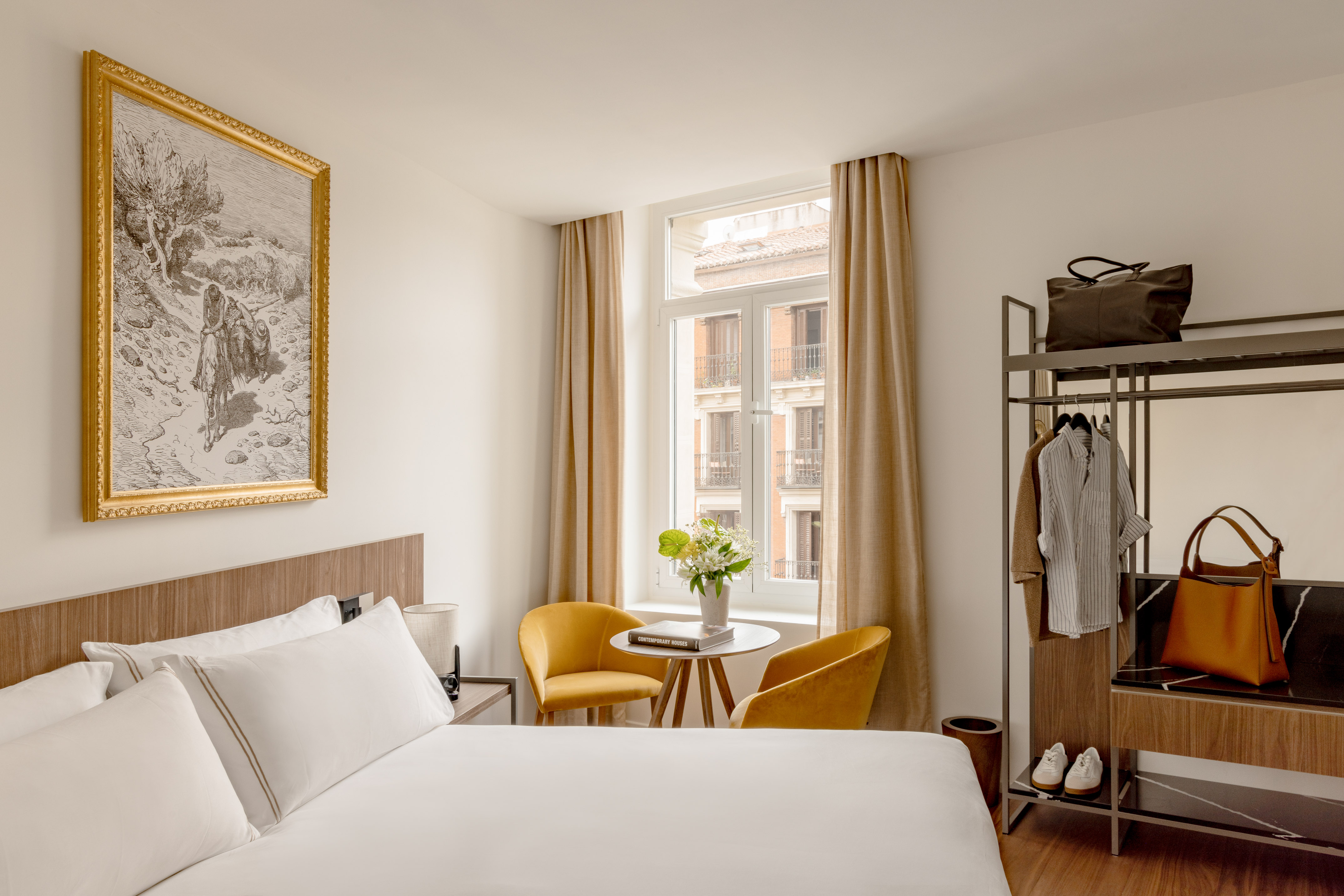 Bright modern hotel bedroom with white bed, window city view, and yellow chairs.