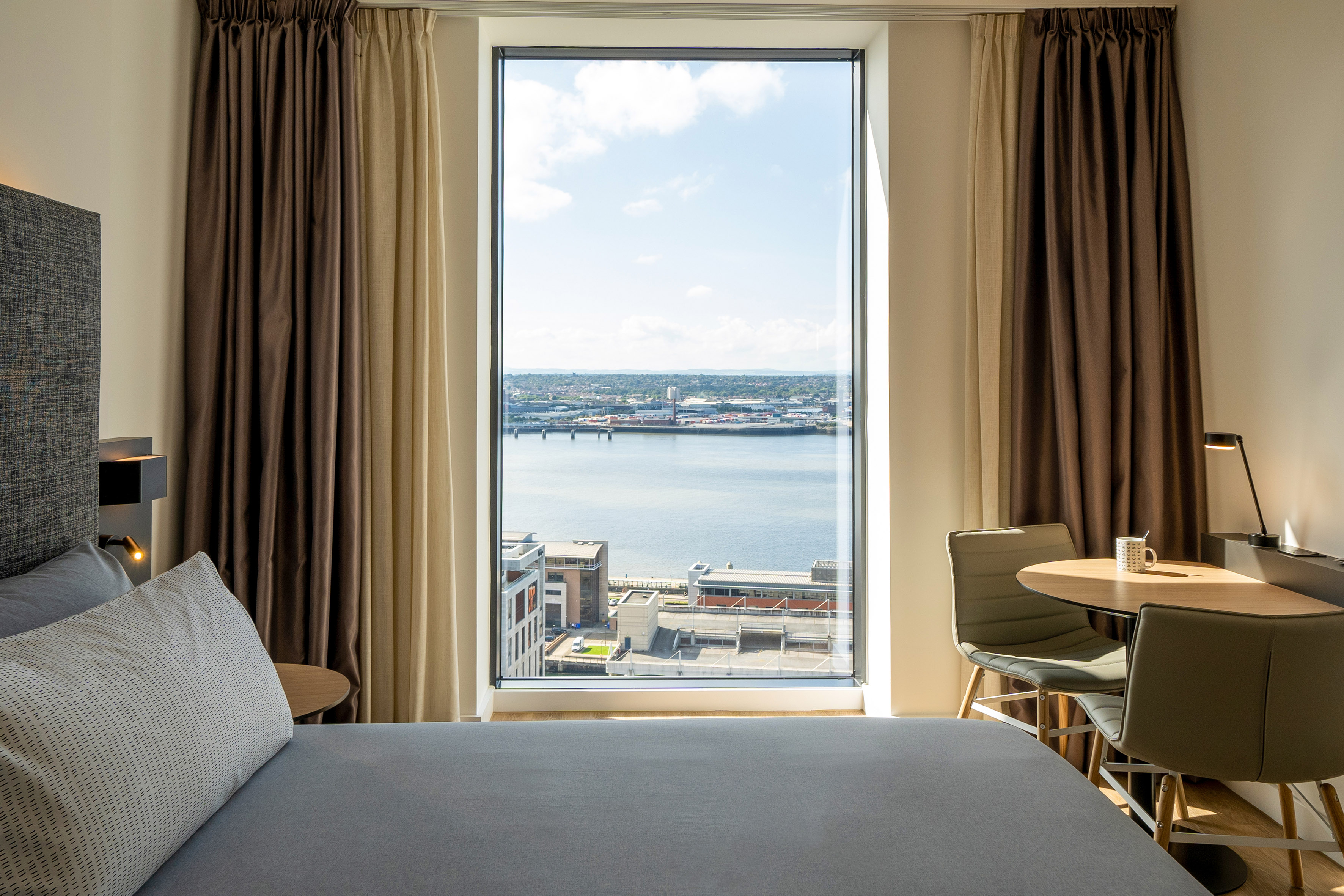 Modern hotel room with large window, city & harbor view, bed, table, chairs.