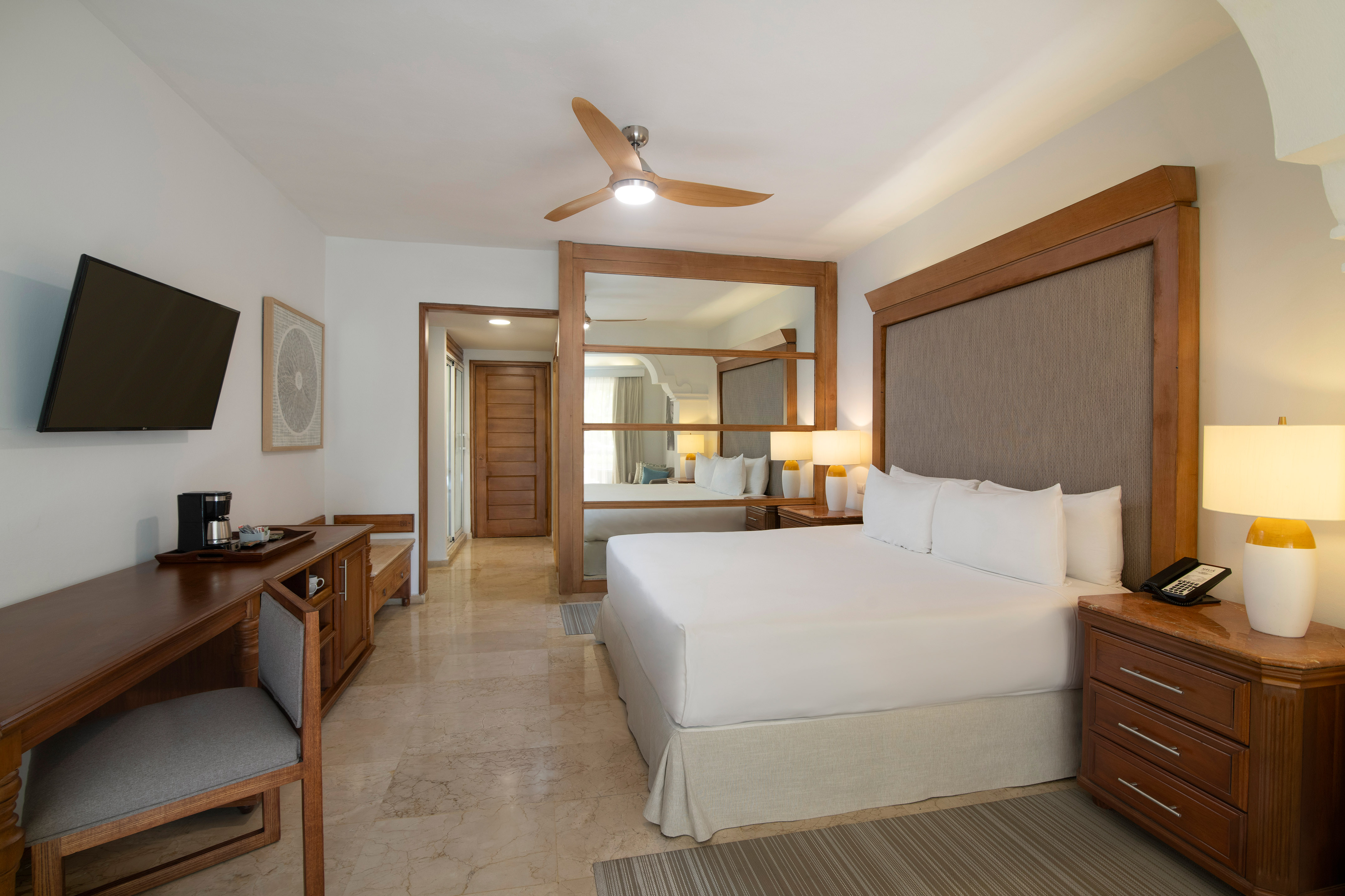 Modern hotel room with king bed, wooden furniture, desk, TV, and ceiling fan.