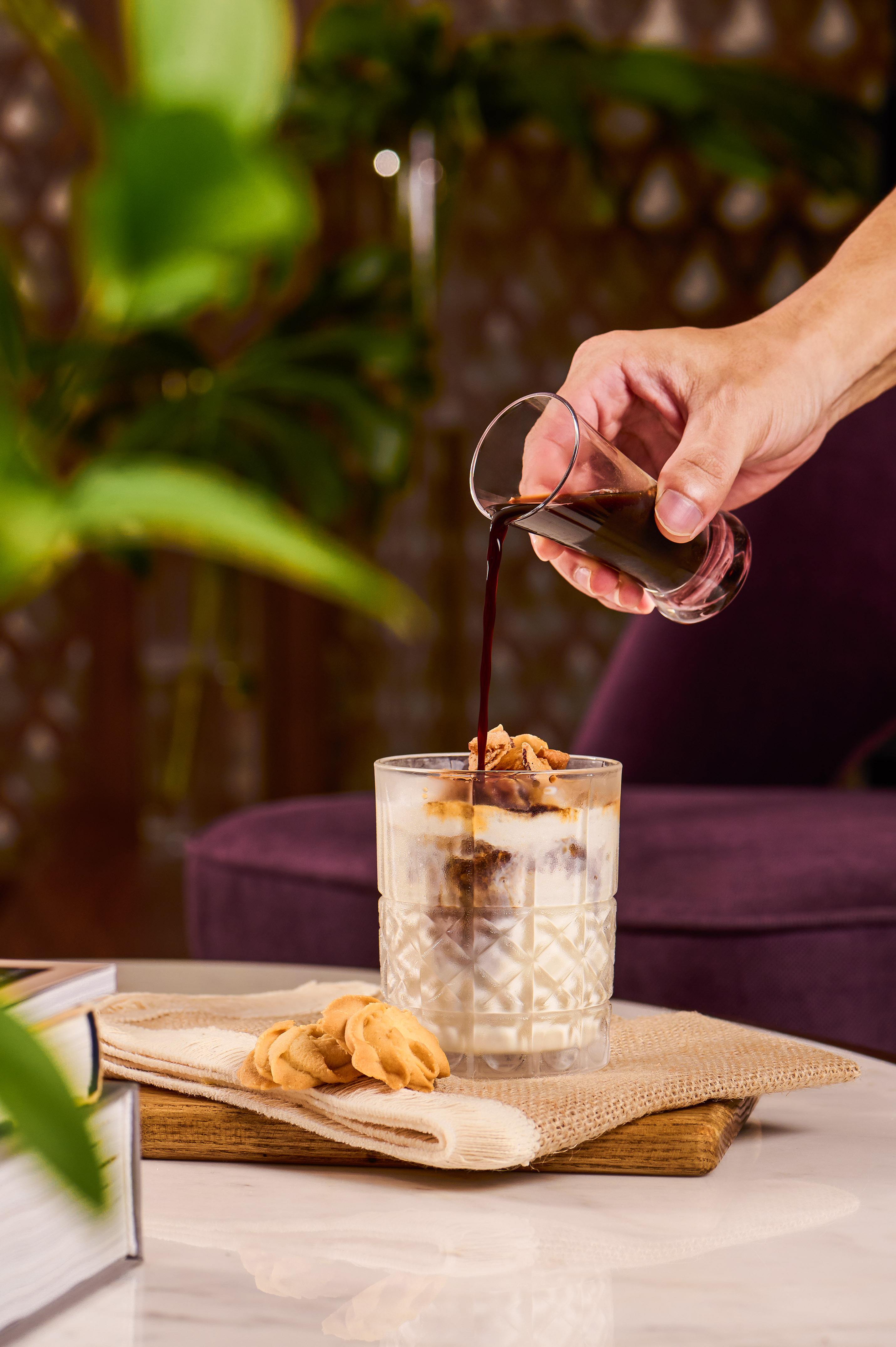 Pouring dark coffee into layered iced latte with cookies on a marble table.