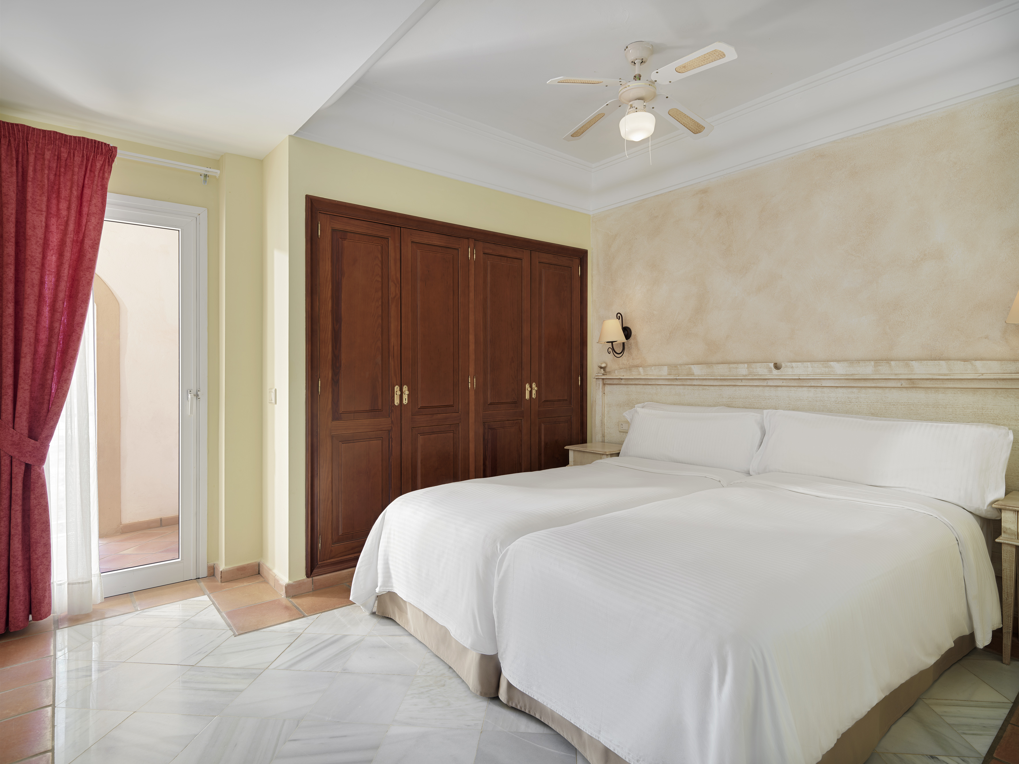 Hotel bedroom with twin beds, wooden wardrobe, ceiling fan, and balcony.