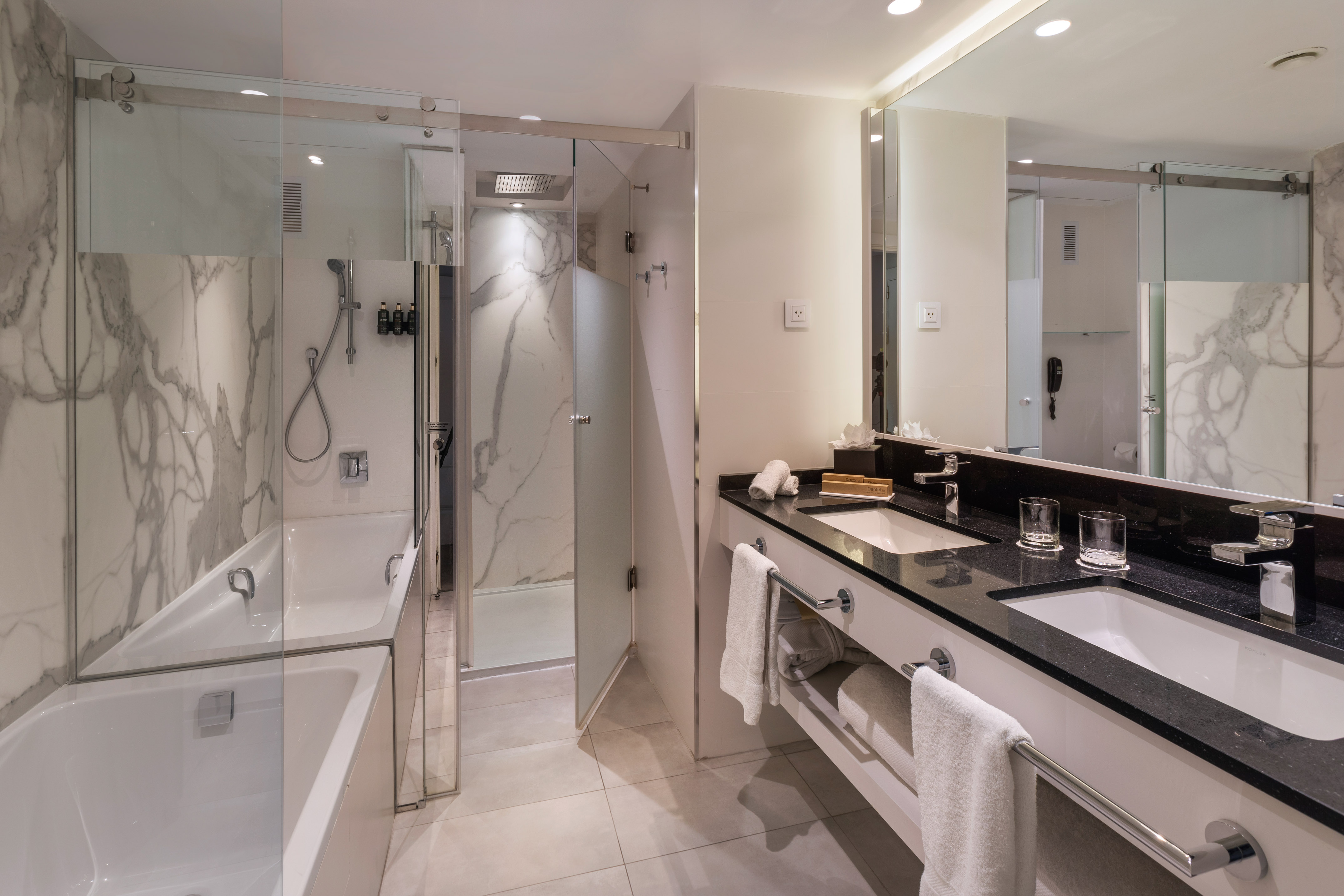 Modern hotel bathroom with bathtub, separate shower, double vanity, and marble walls.