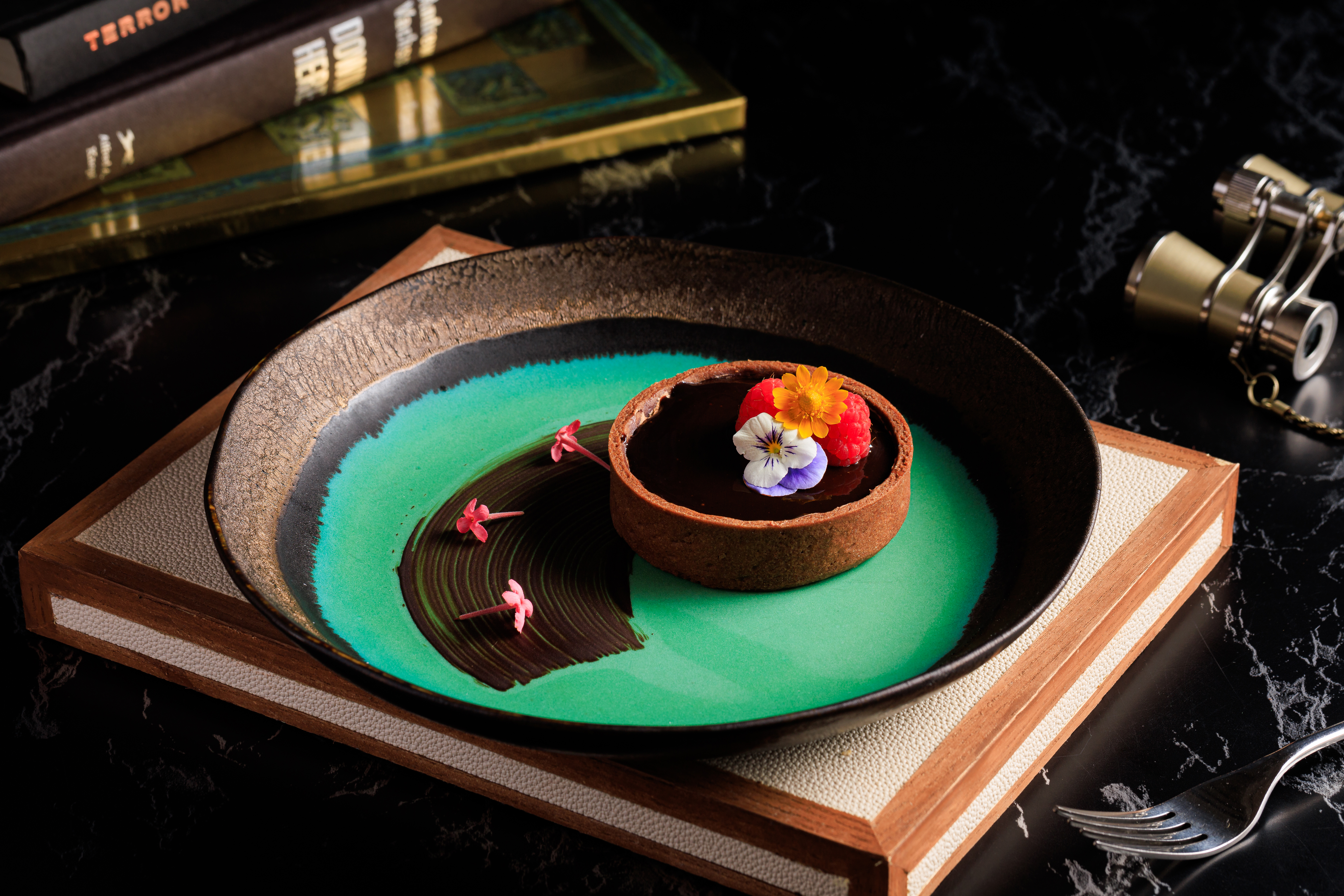 Decadent chocolate tart with edible flowers, raspberries on vibrant green plate, dark setting.