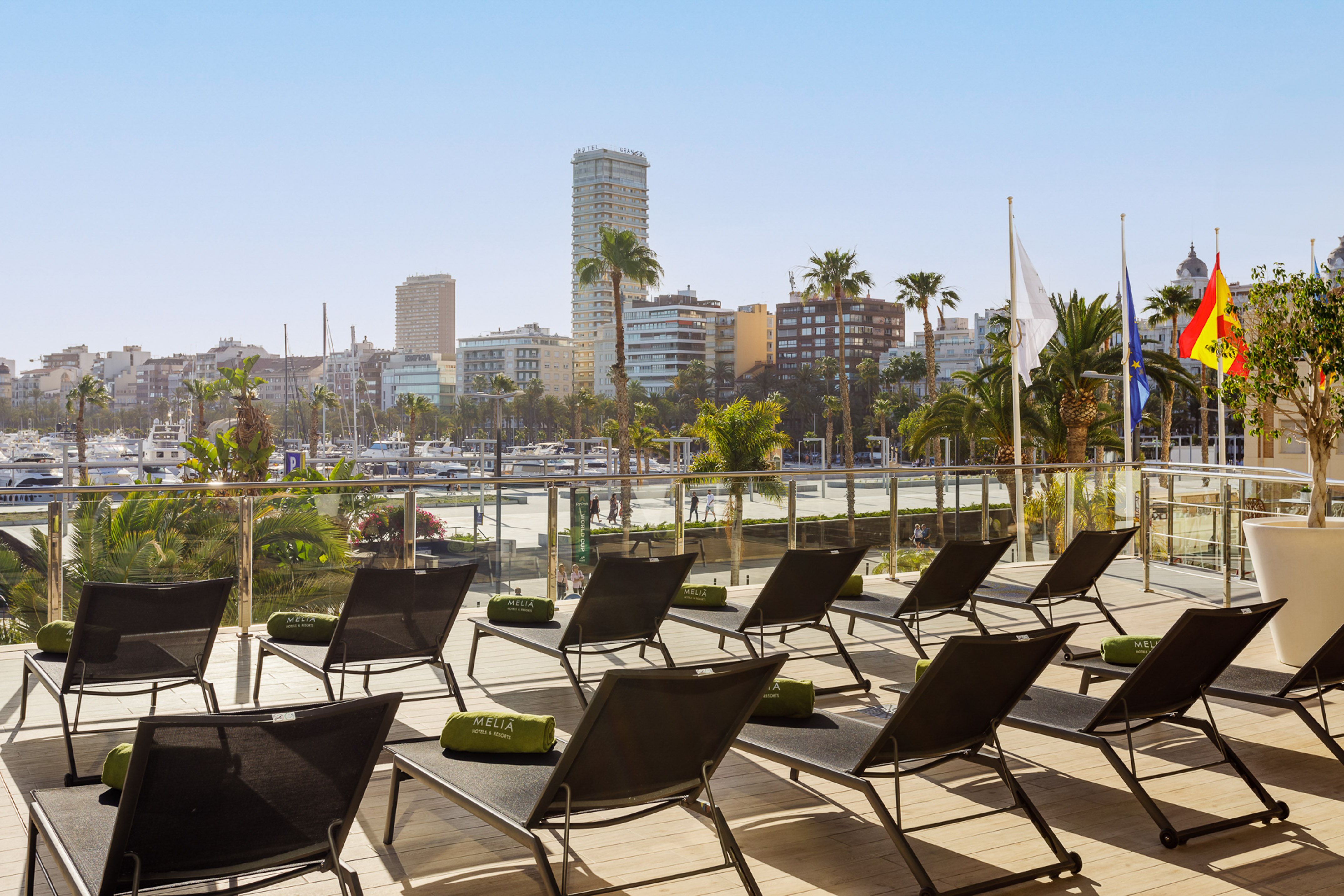 Hotel terrace with sun loungers overlooking marina, city, and palm trees.