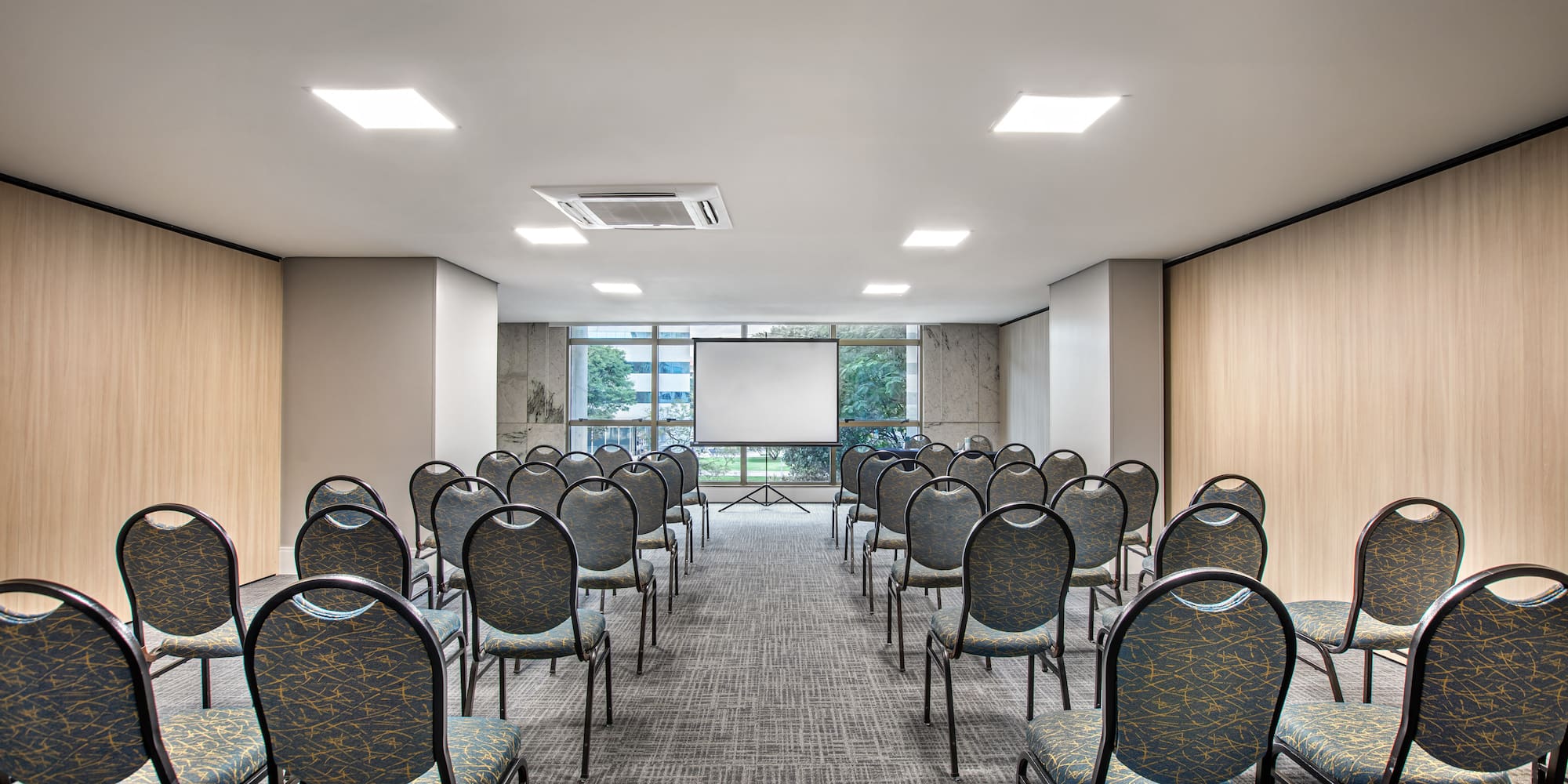 Modern meeting room with rows of chairs, projection screen, and windows.