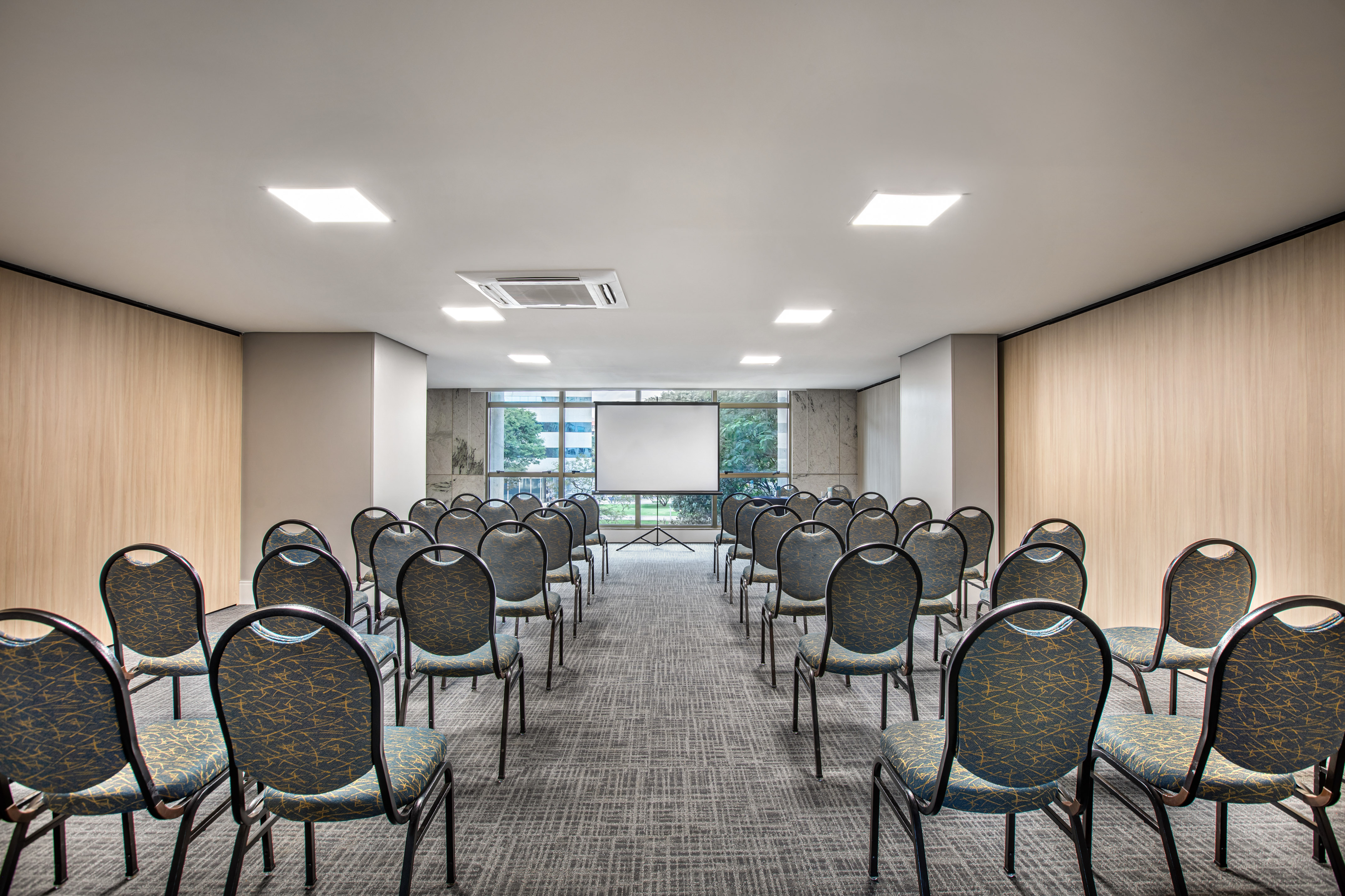 Modern meeting room with rows of chairs, projection screen, and windows.