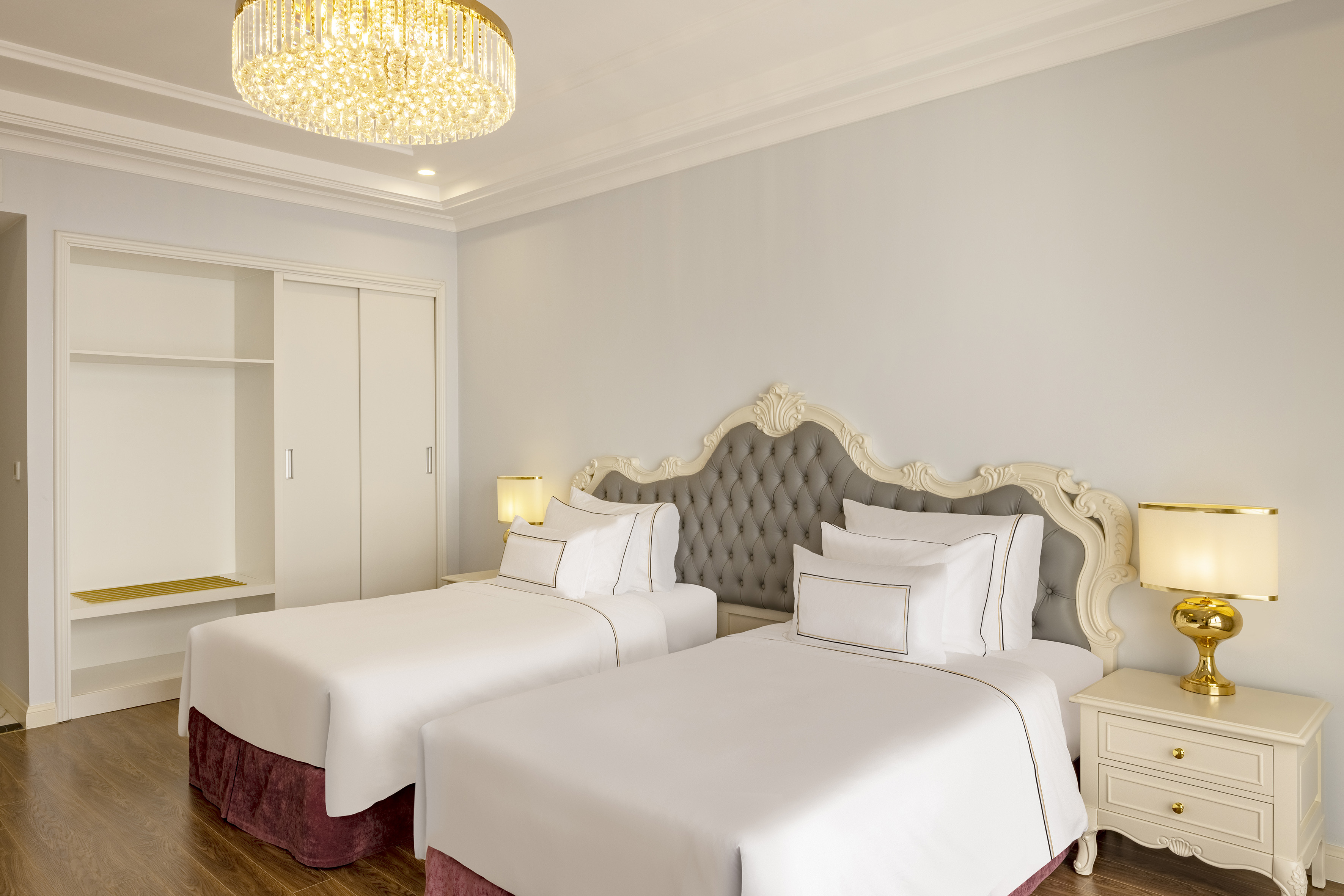 Elegant hotel bedroom with twin beds, ornate gray headboard, and crystal chandelier.