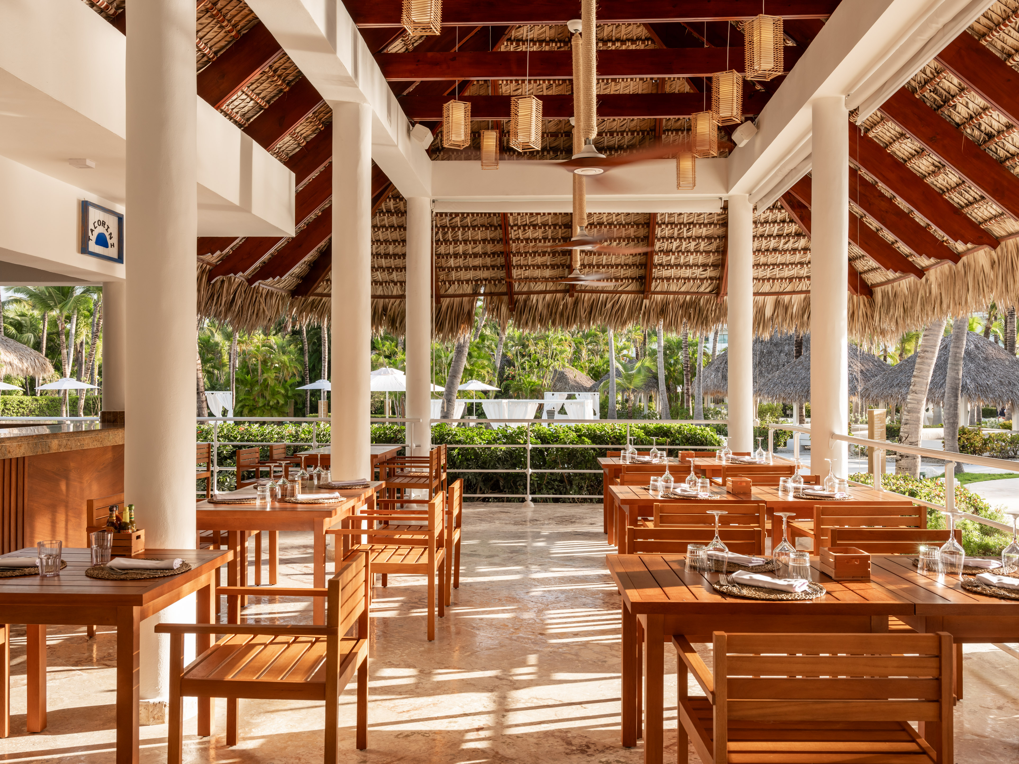 Outdoor resort dining with wooden tables, chairs, and a thatch roof.