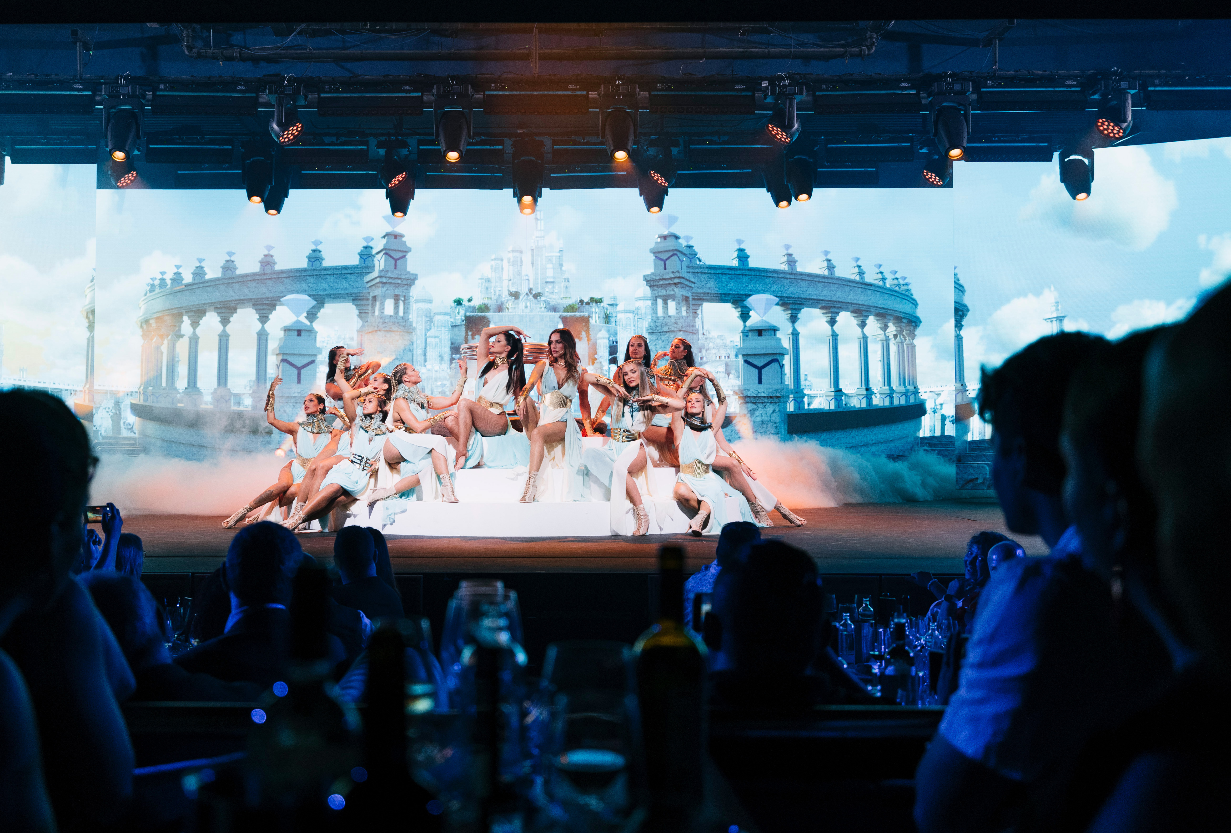 Dancers in elegant ancient-style costumes on stage with a grand palace backdrop.