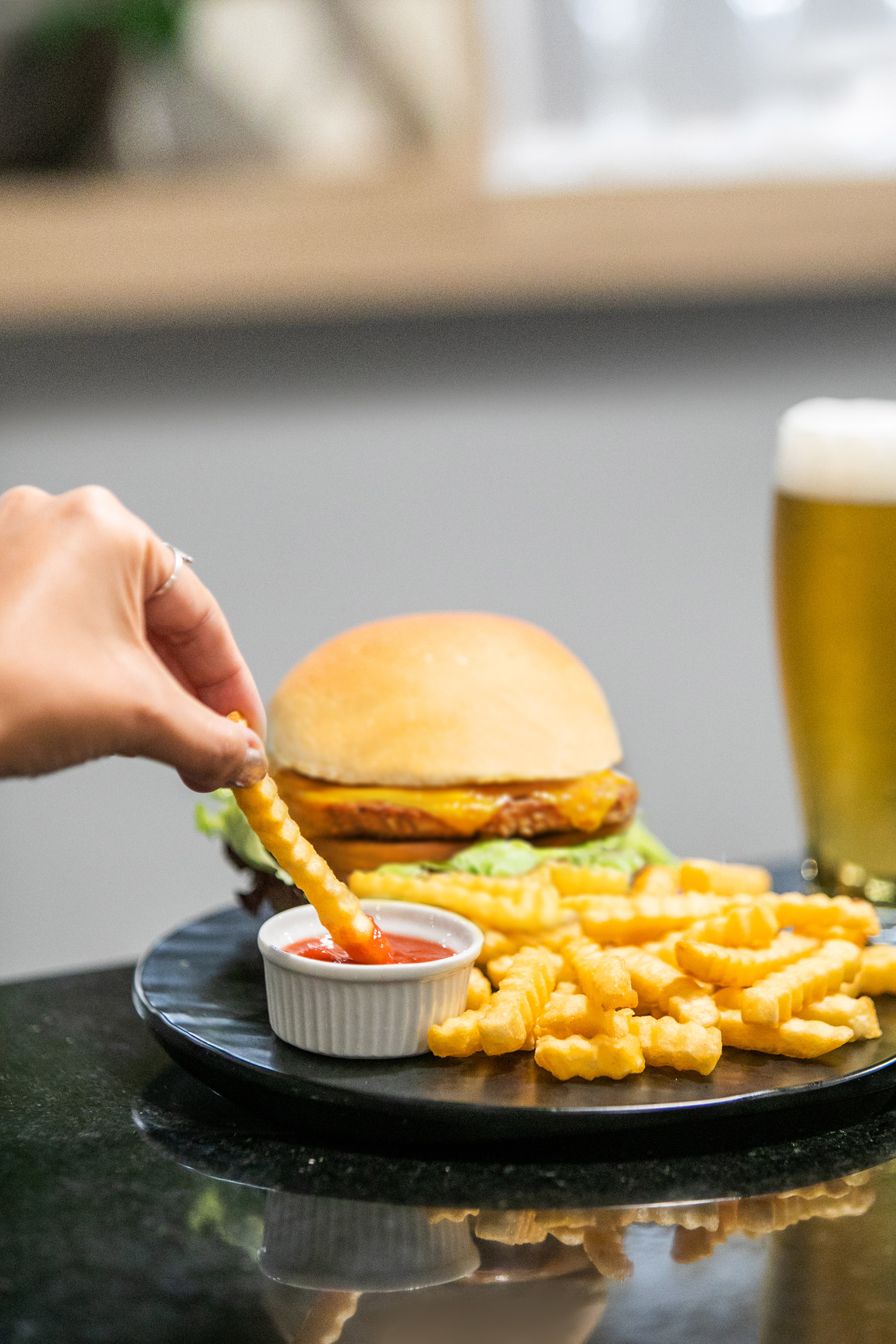 Hand dipping crinkle-cut fry into ketchup, with burger, fries, and beer.