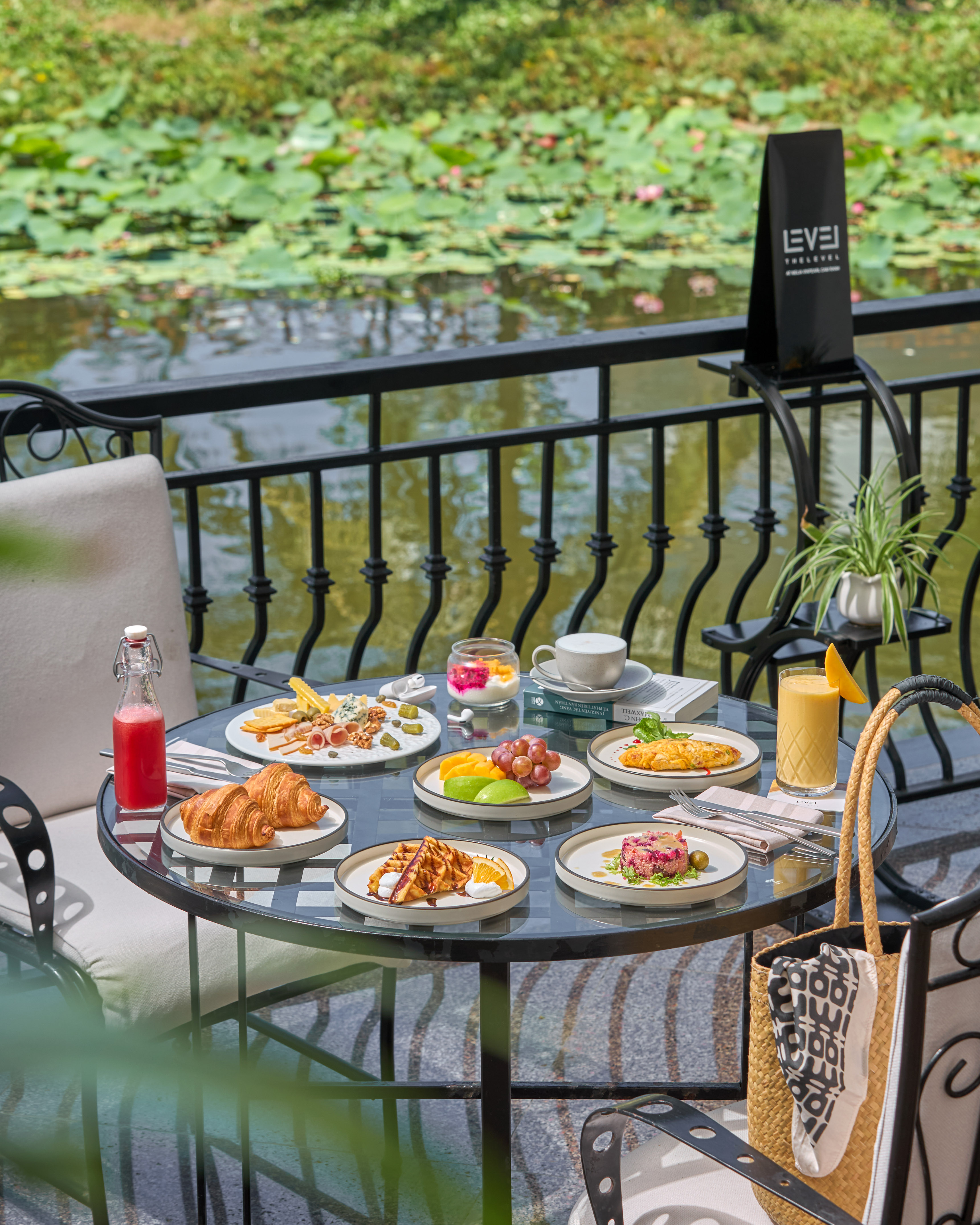 Outdoor breakfast spread with croissants, omelette, fruits, and juices by a lotus pond.