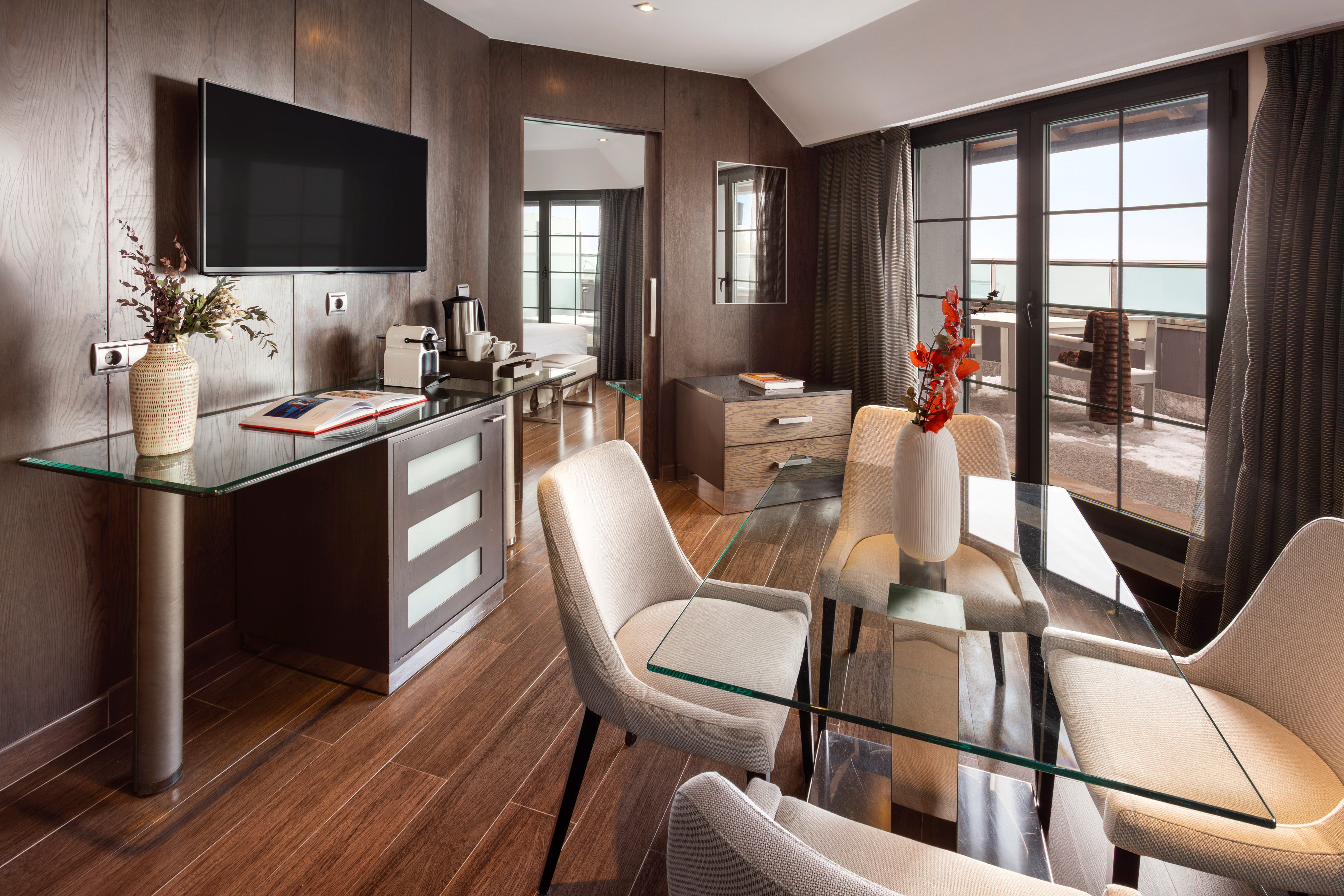 Modern hotel suite living room with dining table, TV, coffee station, balcony.