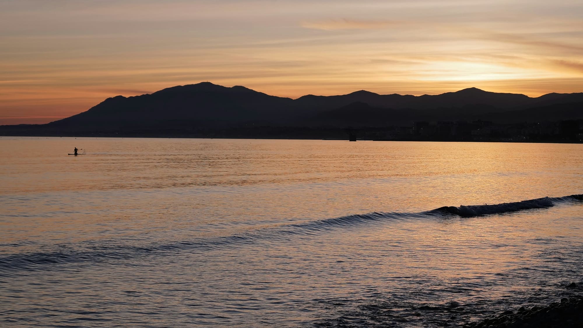 Serene sunset over ocean with paddleboarder, mountains, and gentle waves.