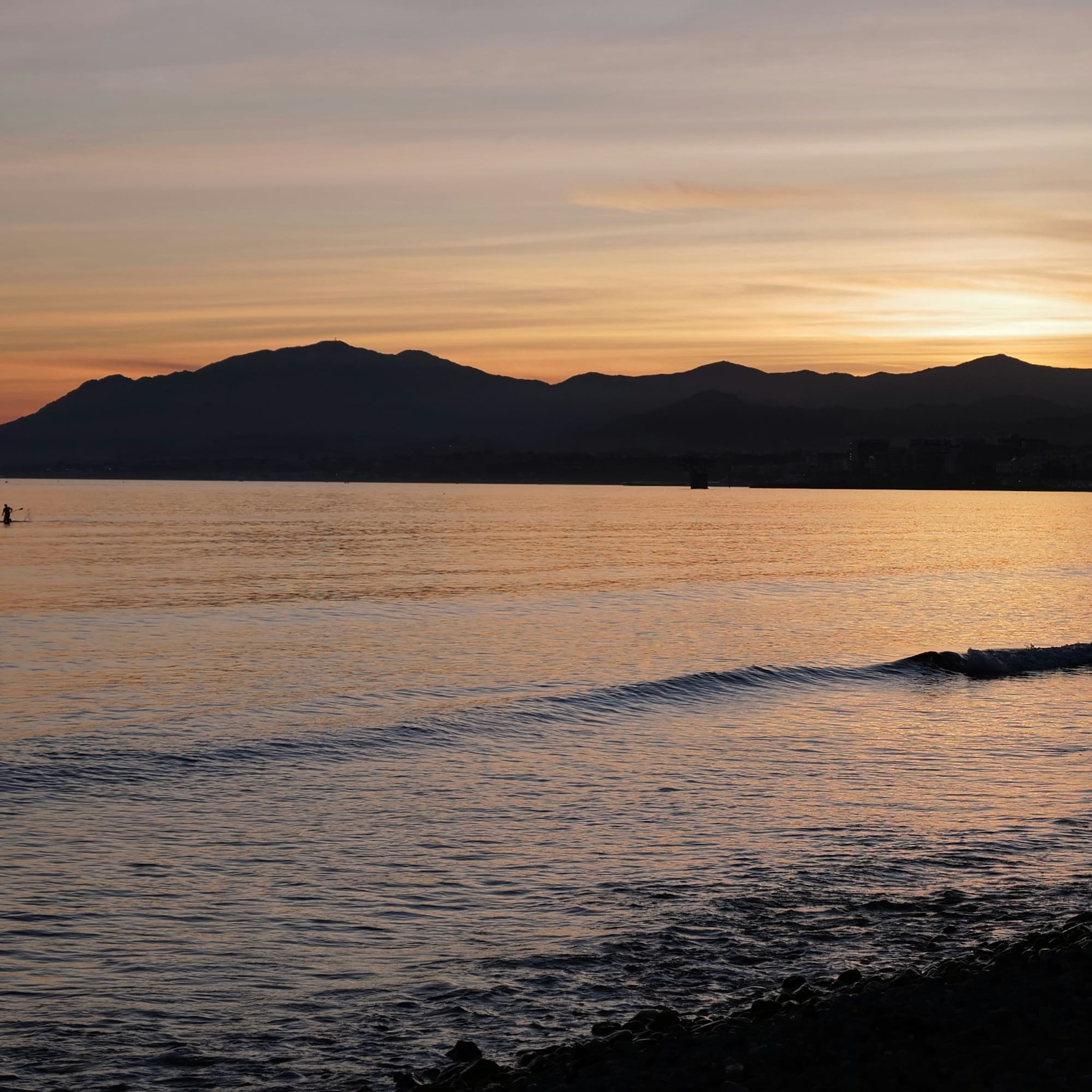 Paddleboarder on ocean at sunset, mountains silhouetted against golden sky.
