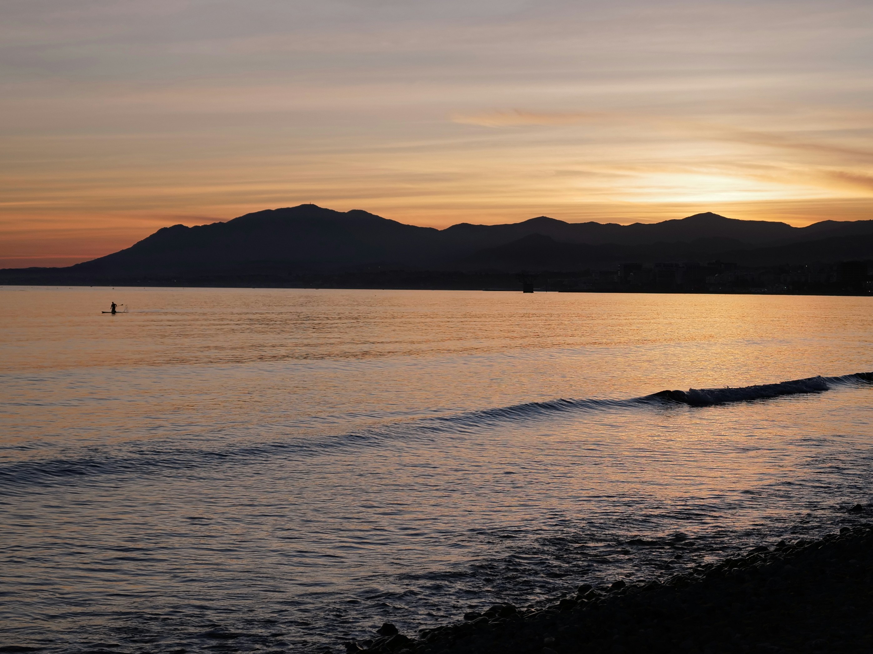Paddleboarder on ocean at sunset, mountains silhouetted against golden sky.
