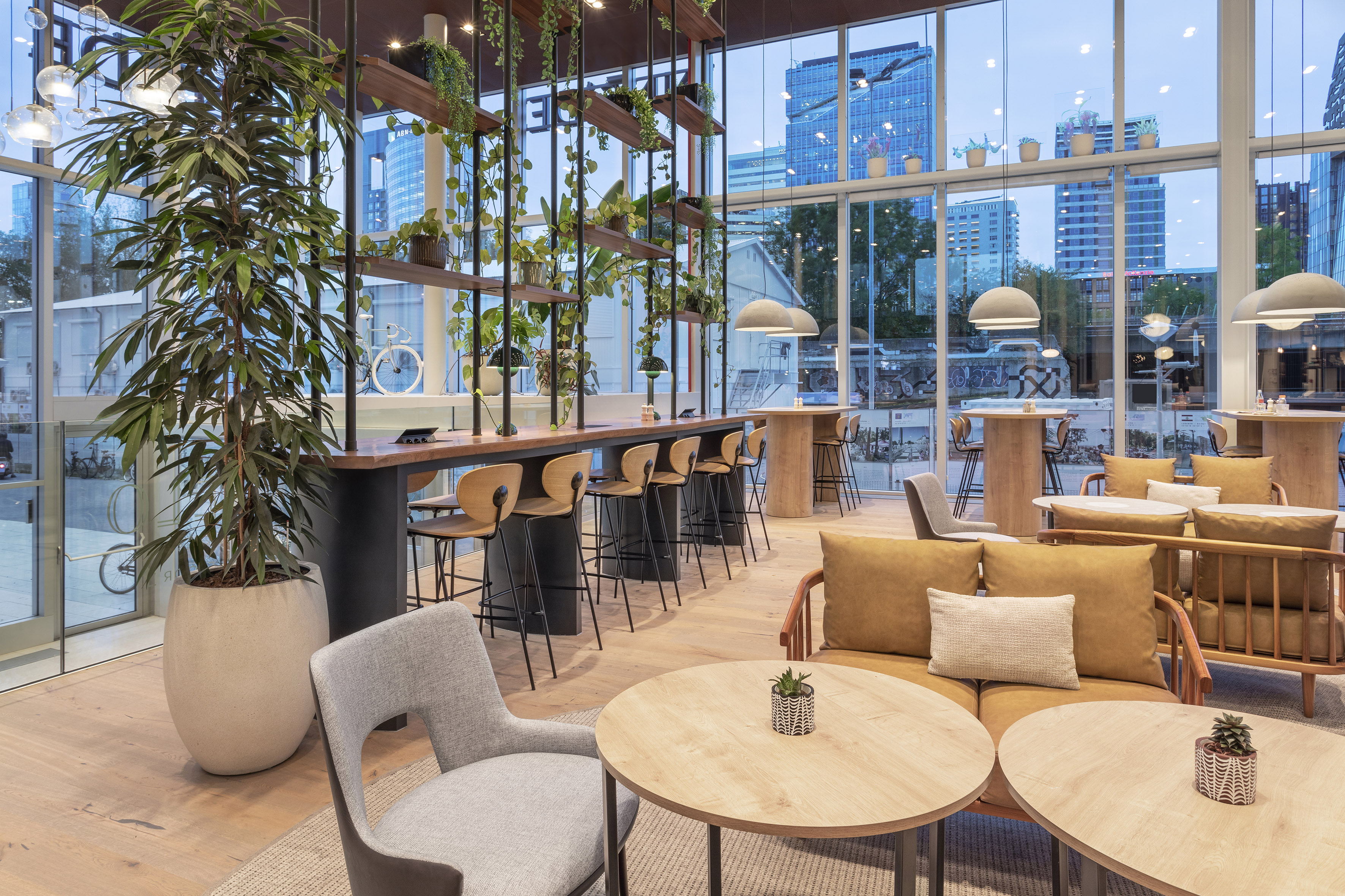 Bright modern lounge with plants, wood bar, diverse seating, and city view.