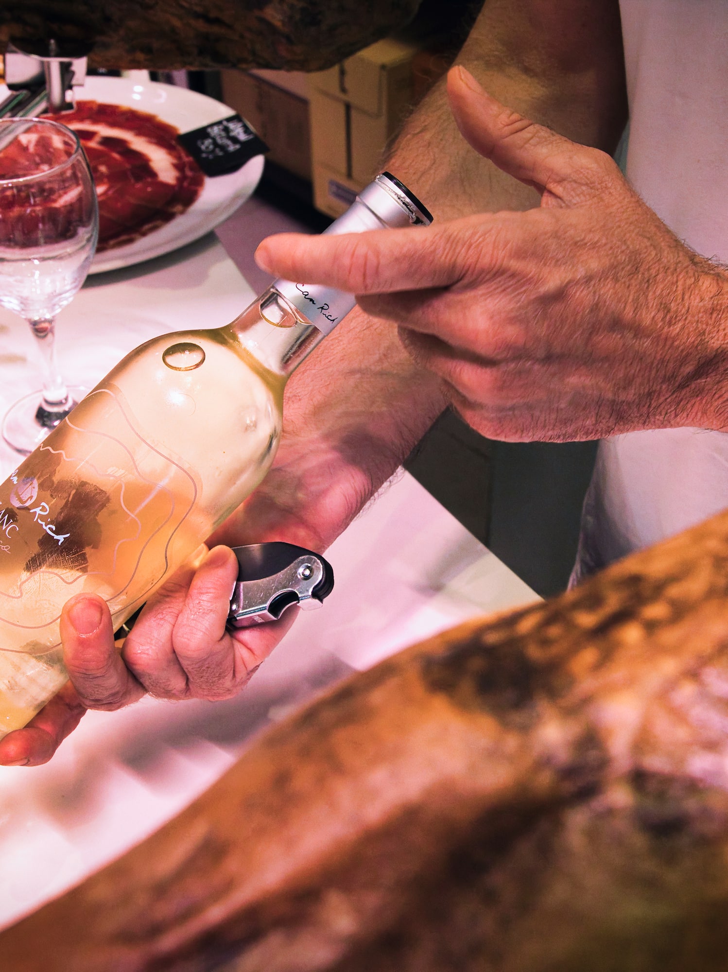 Hands opening "Blanc Rich" white wine bottle next to jamón and glasses.