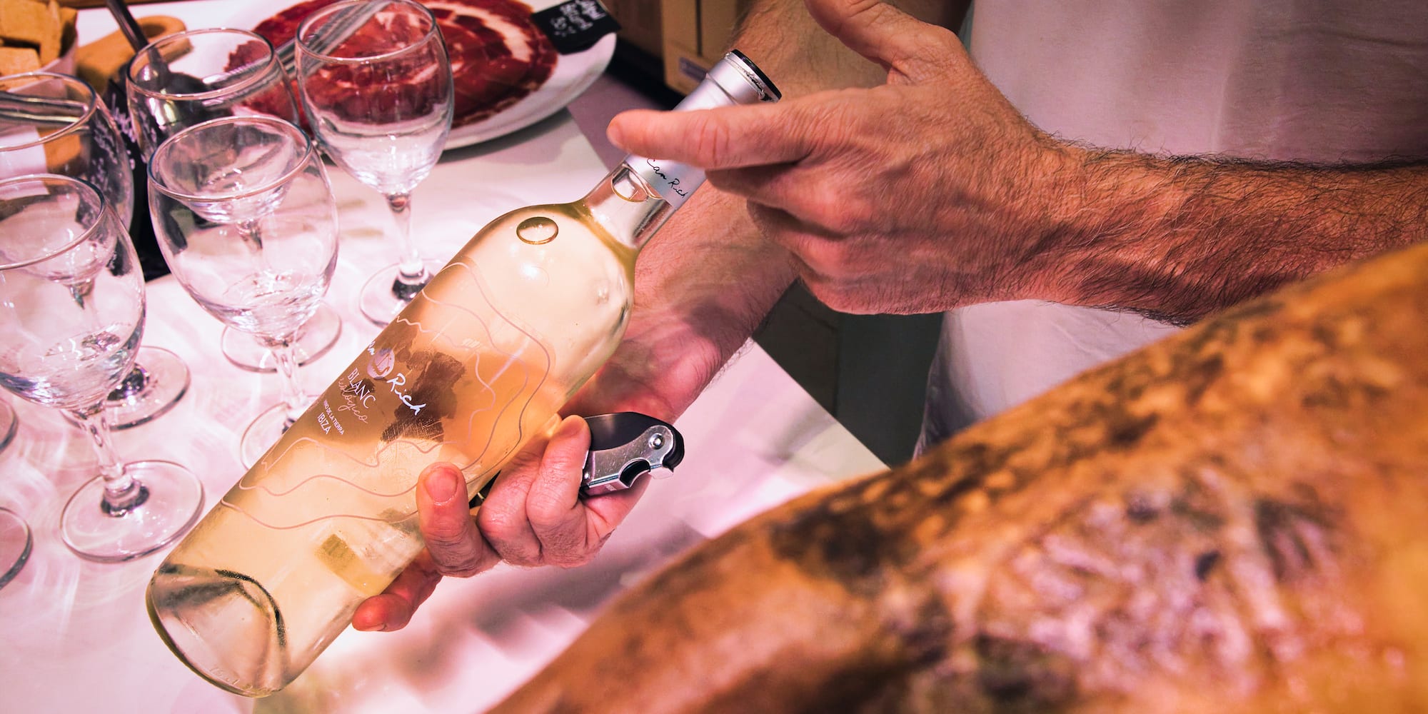Hands opening "Blanc Rich" white wine bottle next to jamón and glasses.
