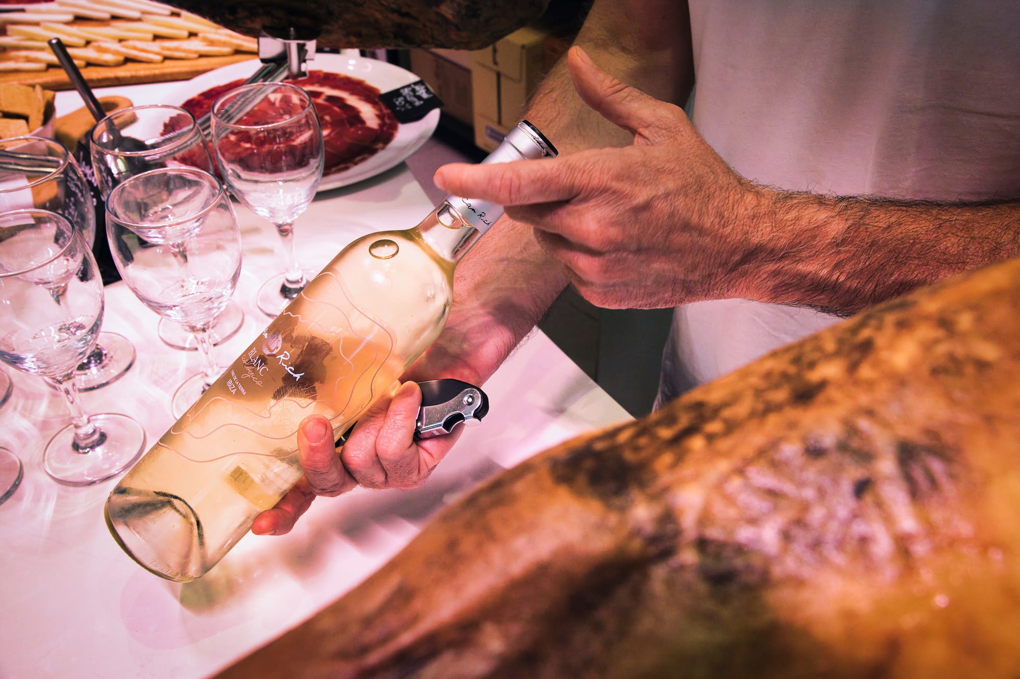 Hands opening "Blanc Rich" white wine bottle next to jamón and glasses.
