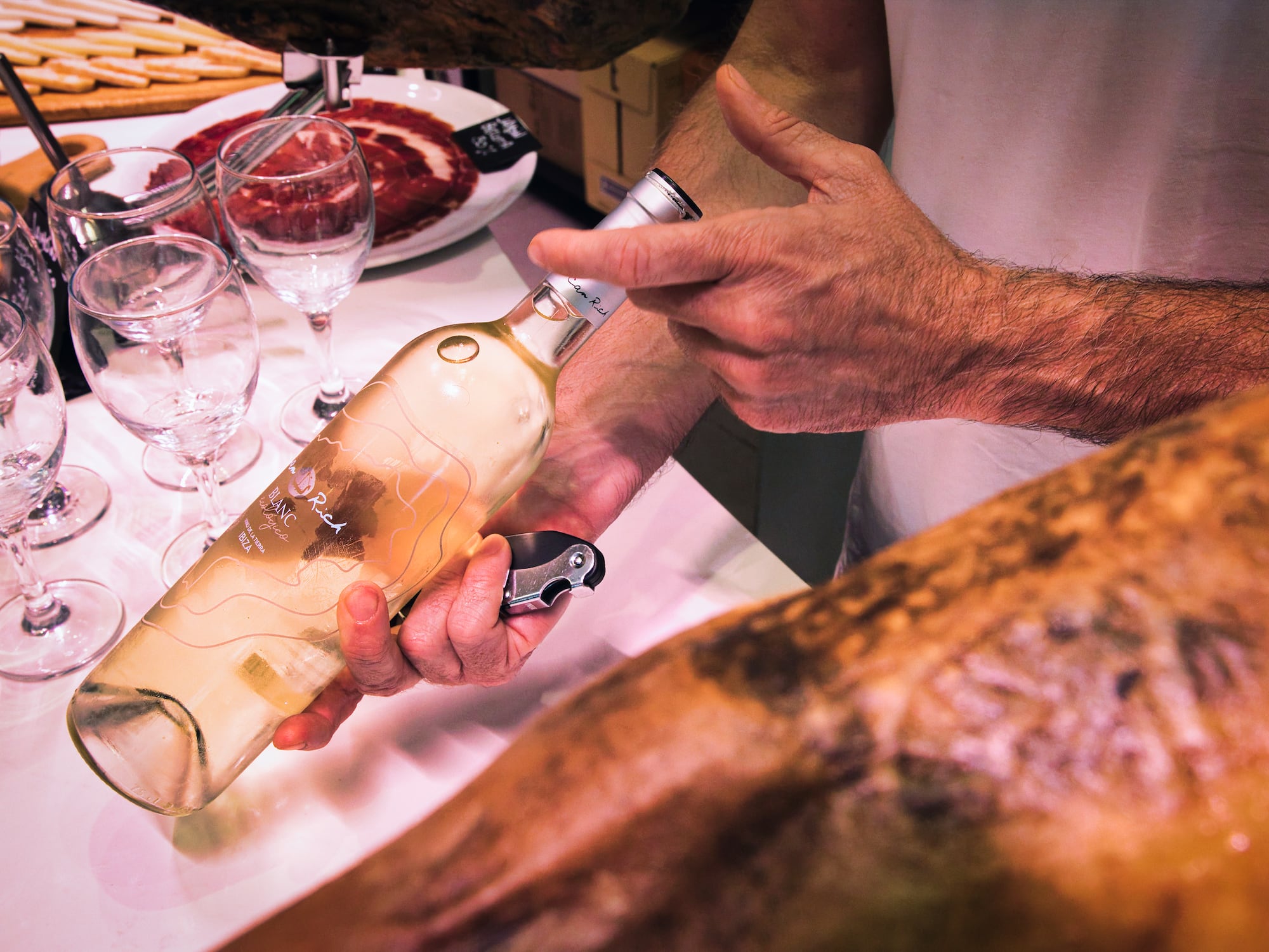 Hands opening "Blanc Rich" white wine bottle next to jamón and glasses.
