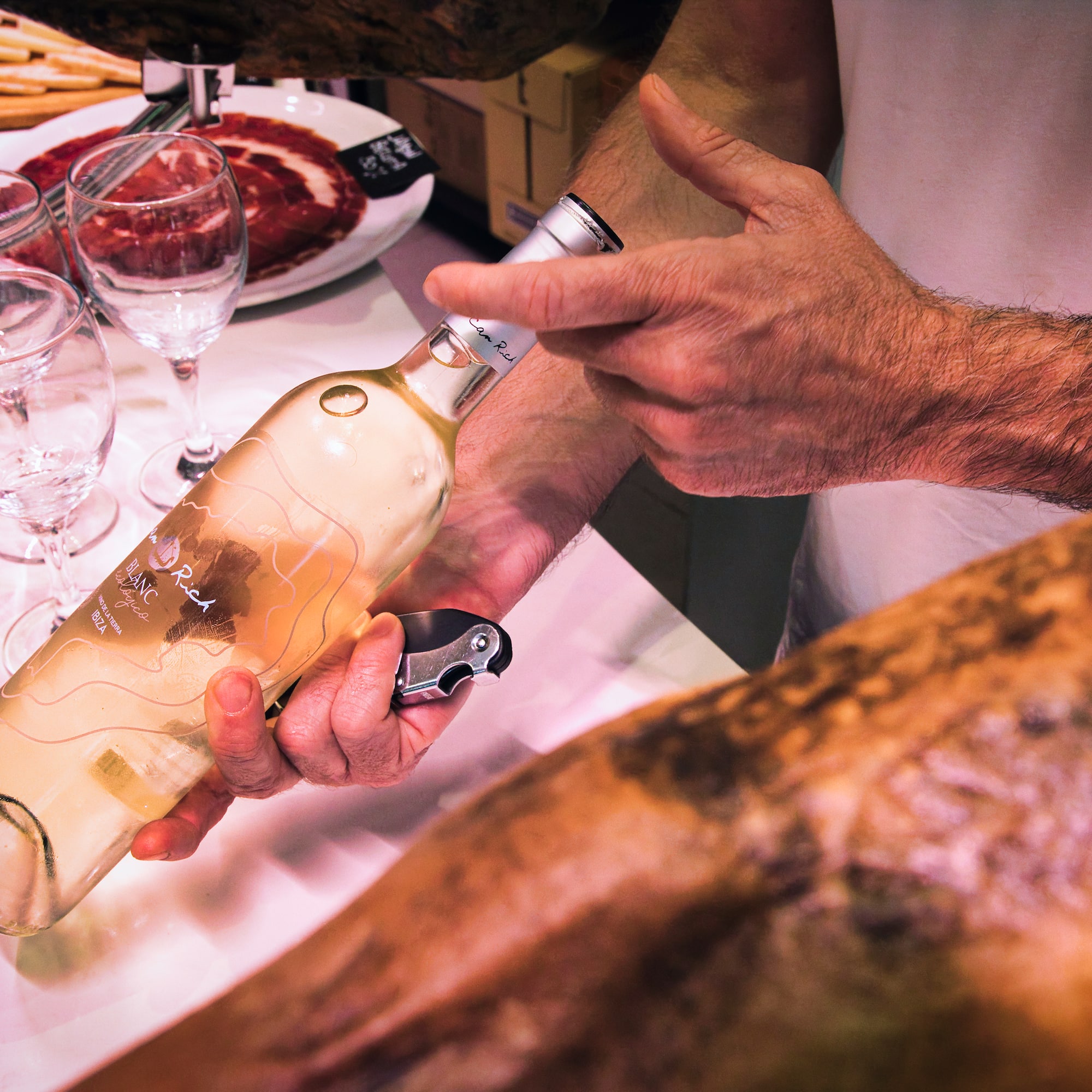 Hands opening "Blanc Rich" white wine bottle next to jamón and glasses.