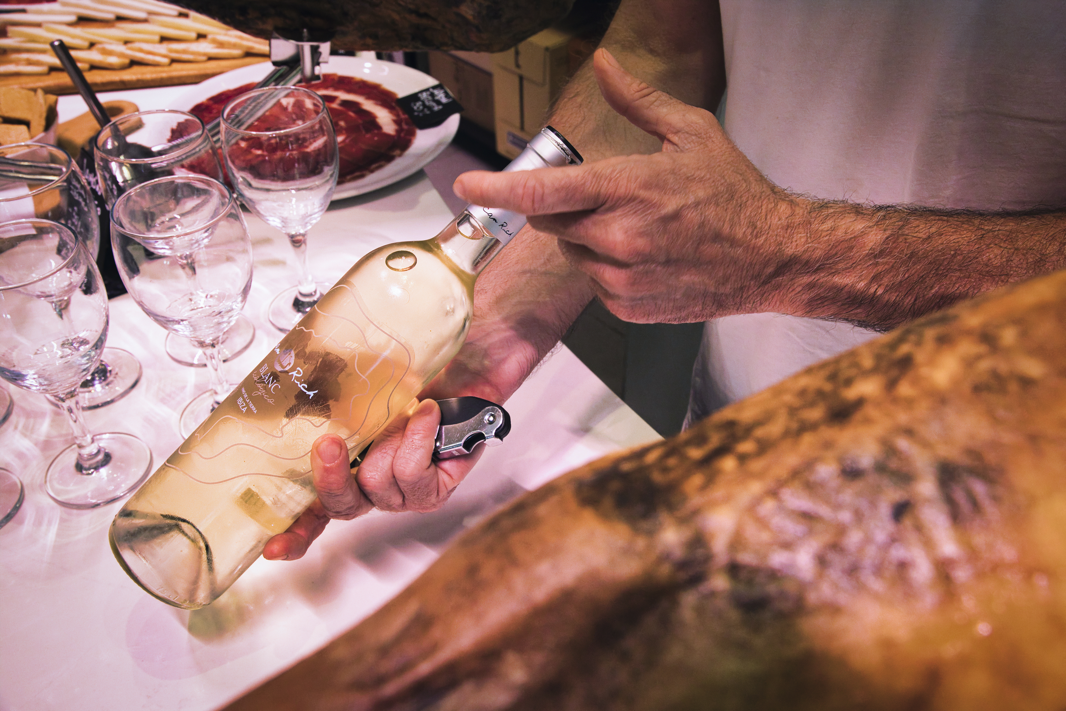 Hands opening "Blanc Rich" white wine bottle next to jamón and glasses.
