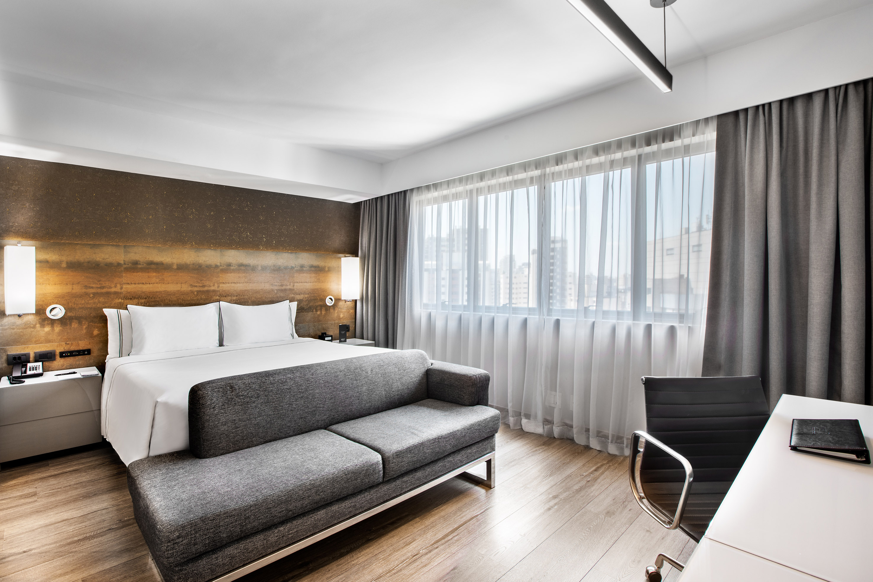Contemporary hotel bedroom with king bed, sofa, workspace, and bright window.
