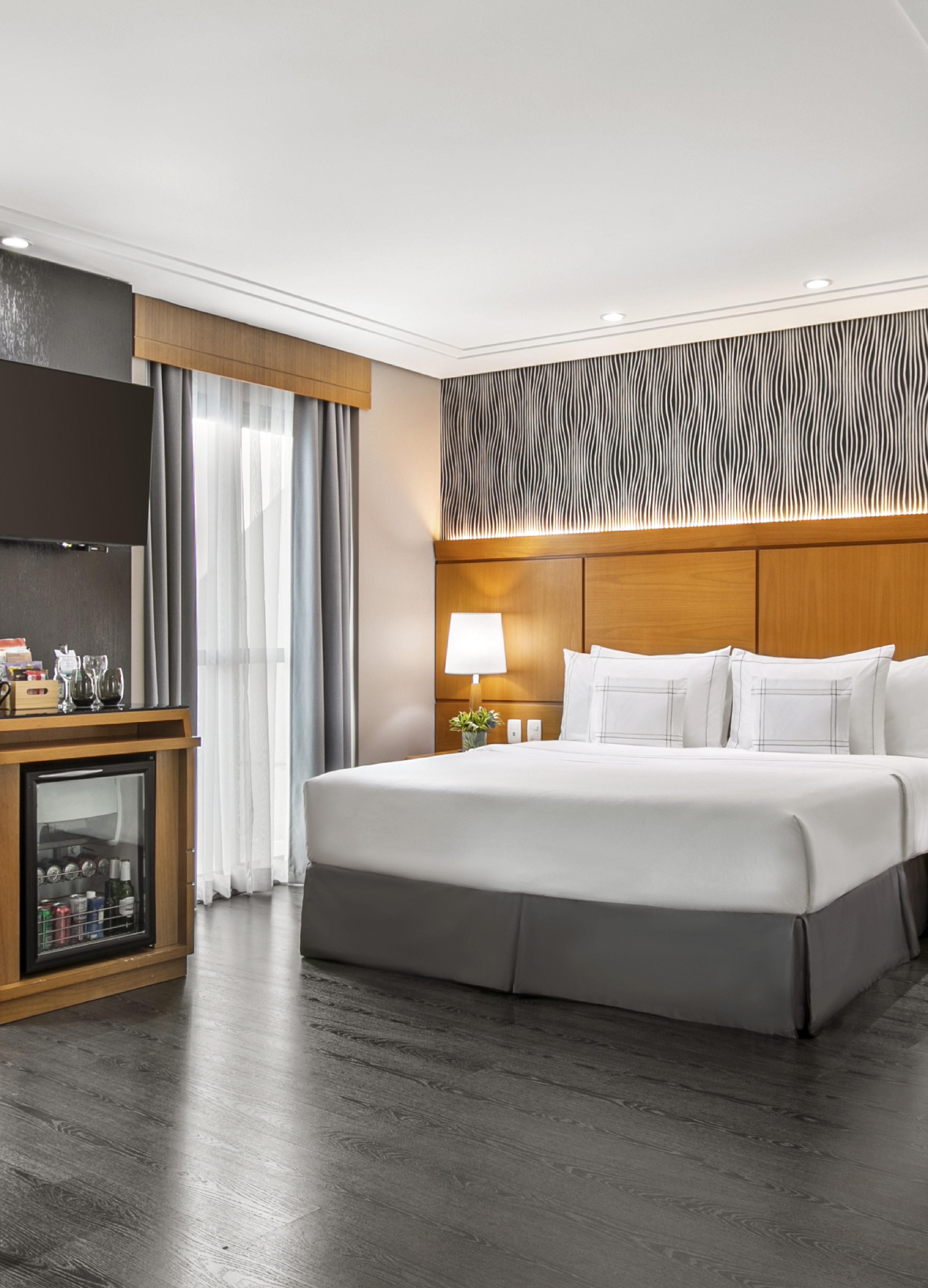 Modern hotel room with king bed, minibar, TV, and dark wood floor.