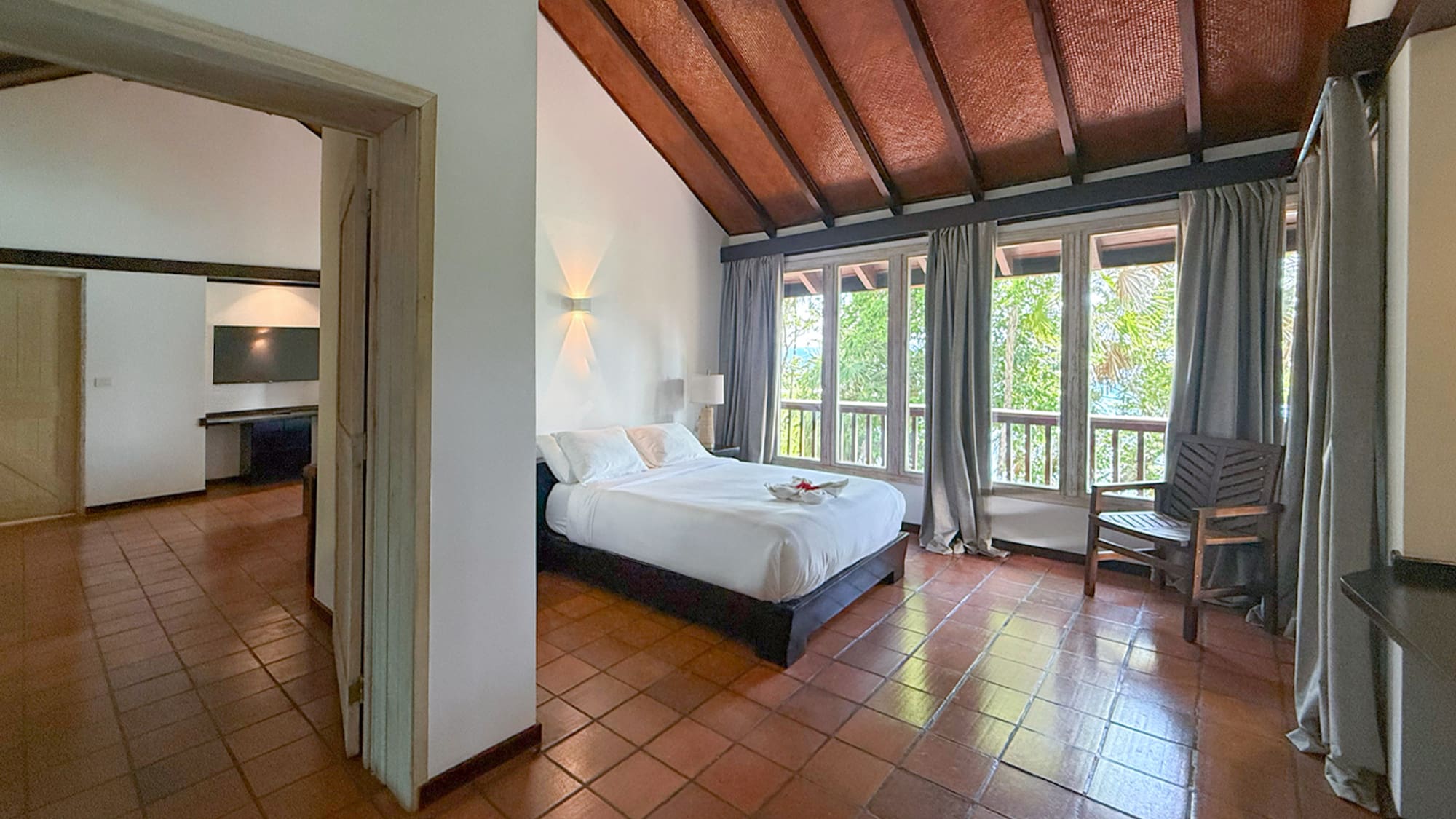 Spacious bedroom with white bed, terracotta floor, vaulted wood ceiling, large windows.