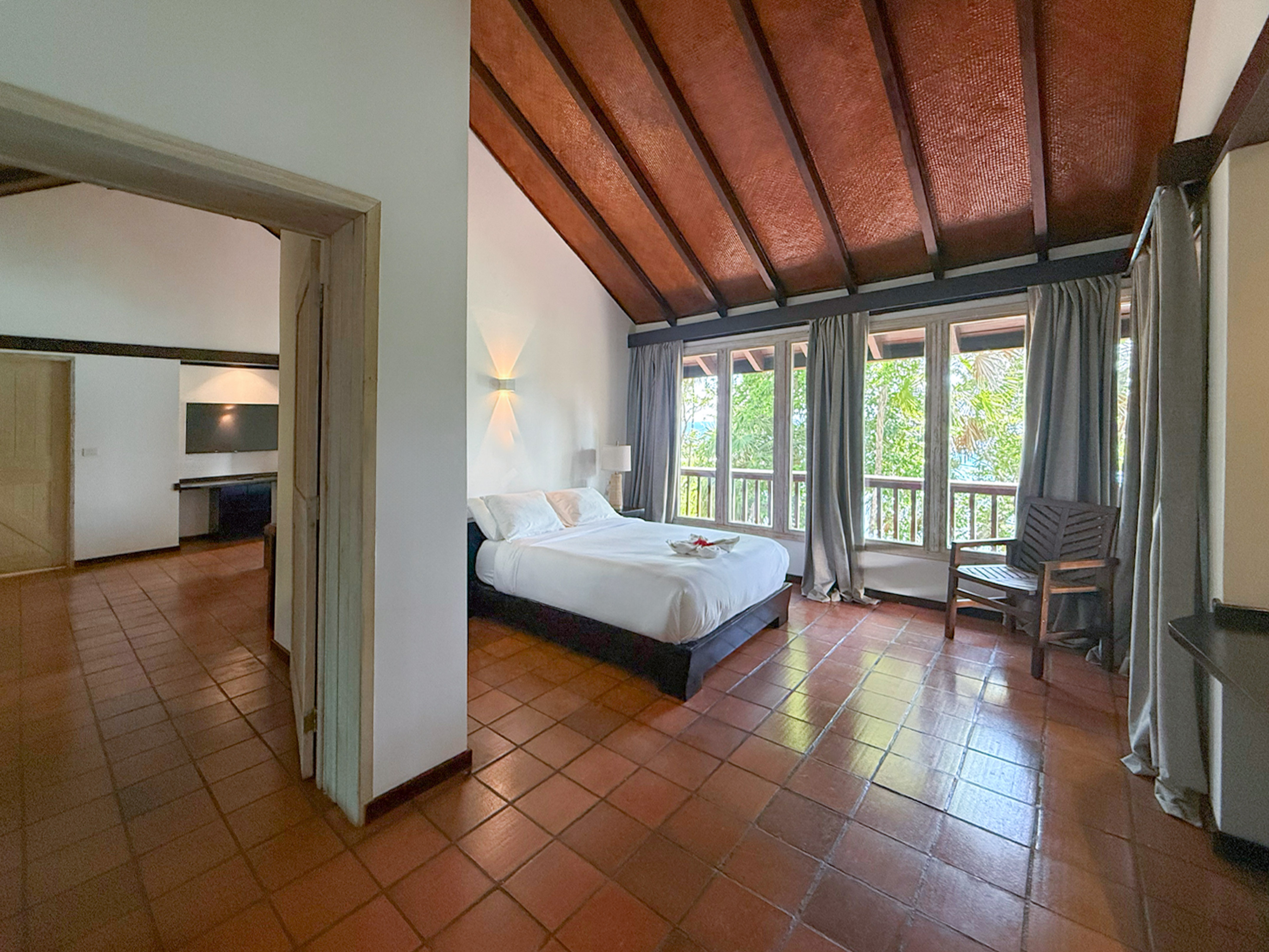 Spacious bedroom with white bed, terracotta floor, vaulted wood ceiling, large windows.