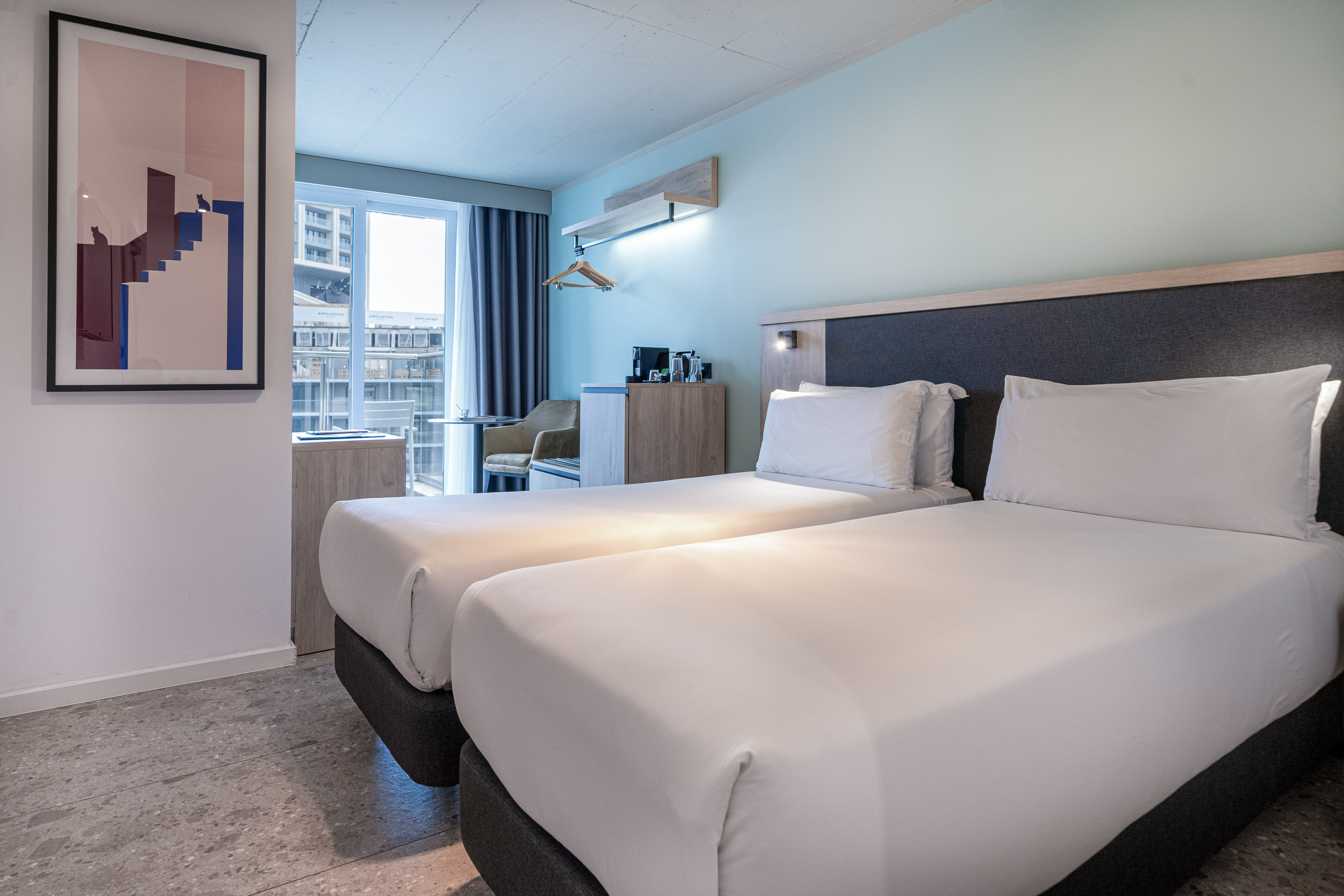 Modern hotel room with two single beds and a city view.