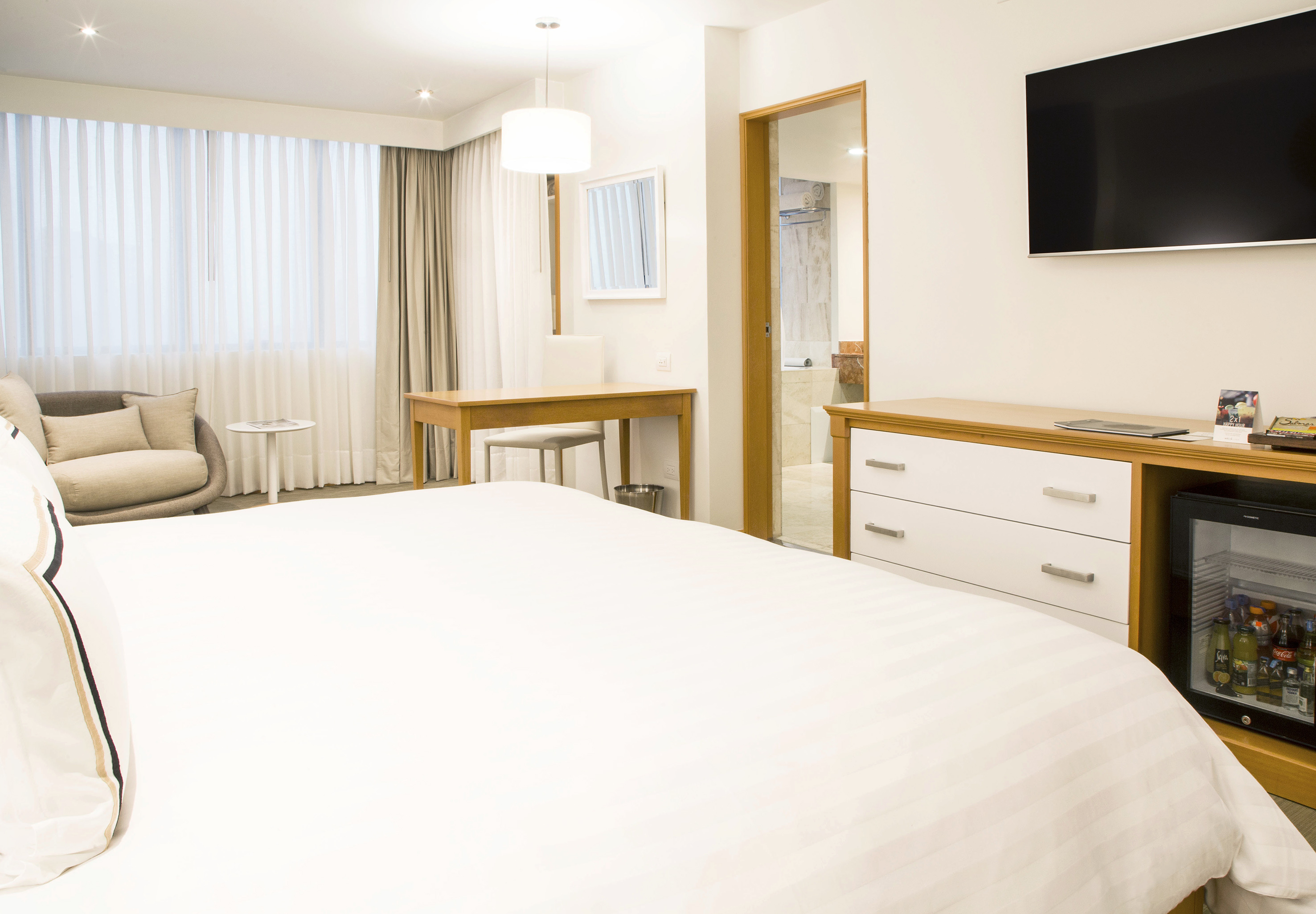 Modern hotel room with large bed, TV, minibar, desk, and window.