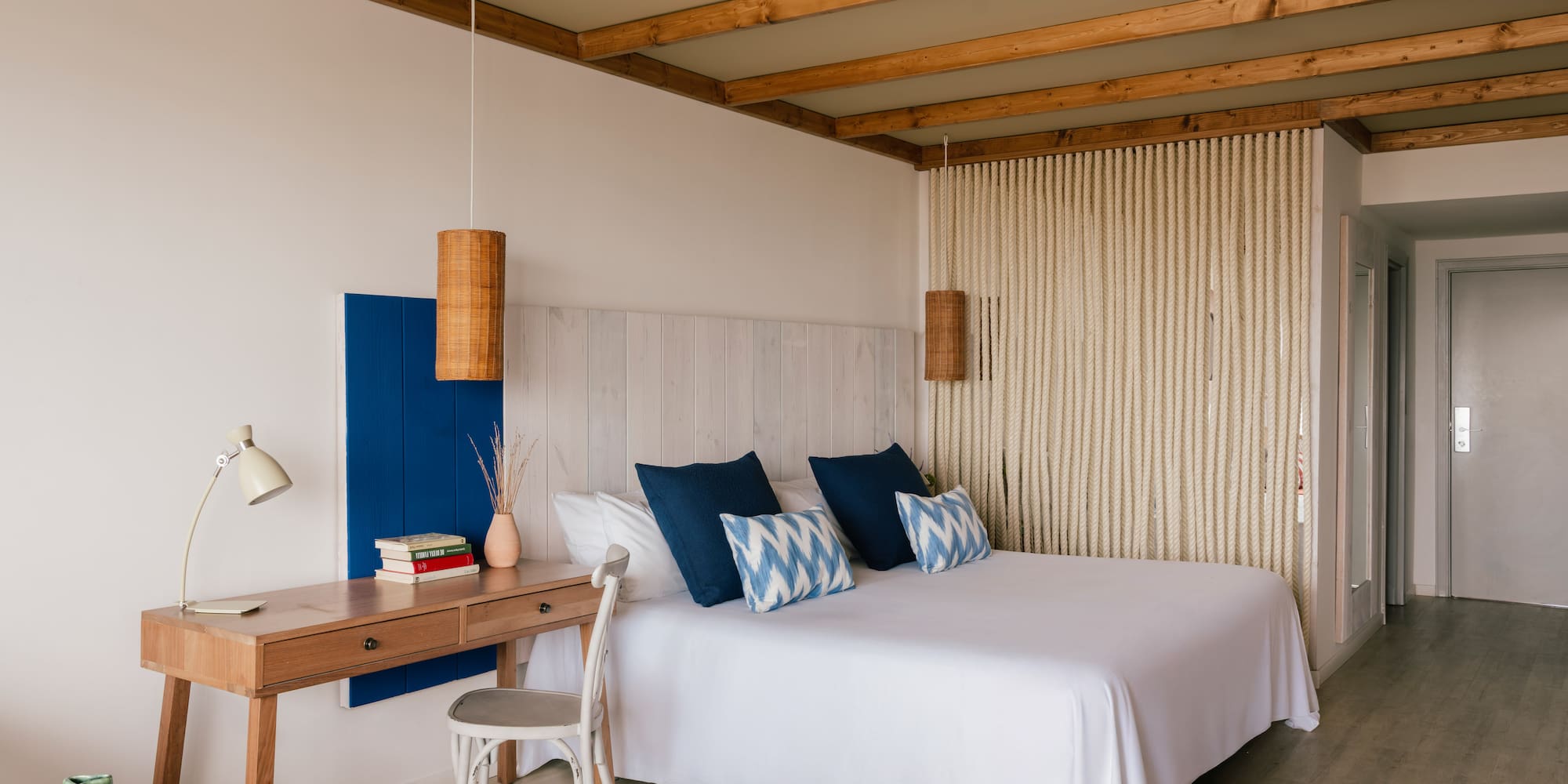 Dormitorio estilo mediterráneo con cama blanca, cojines azules, escritorio y vigas.