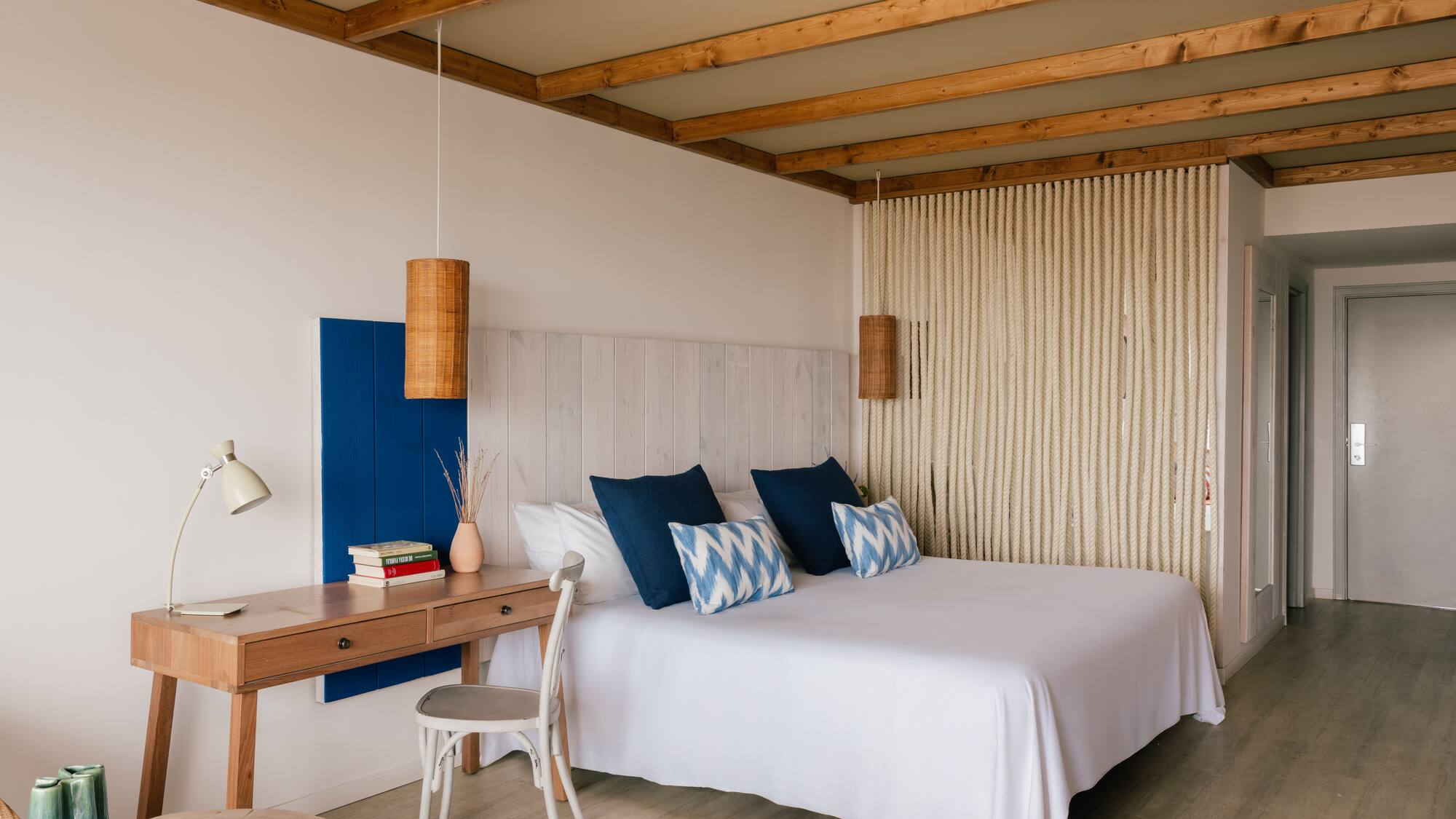 Dormitorio estilo mediterráneo con cama blanca, cojines azules, escritorio y vigas.