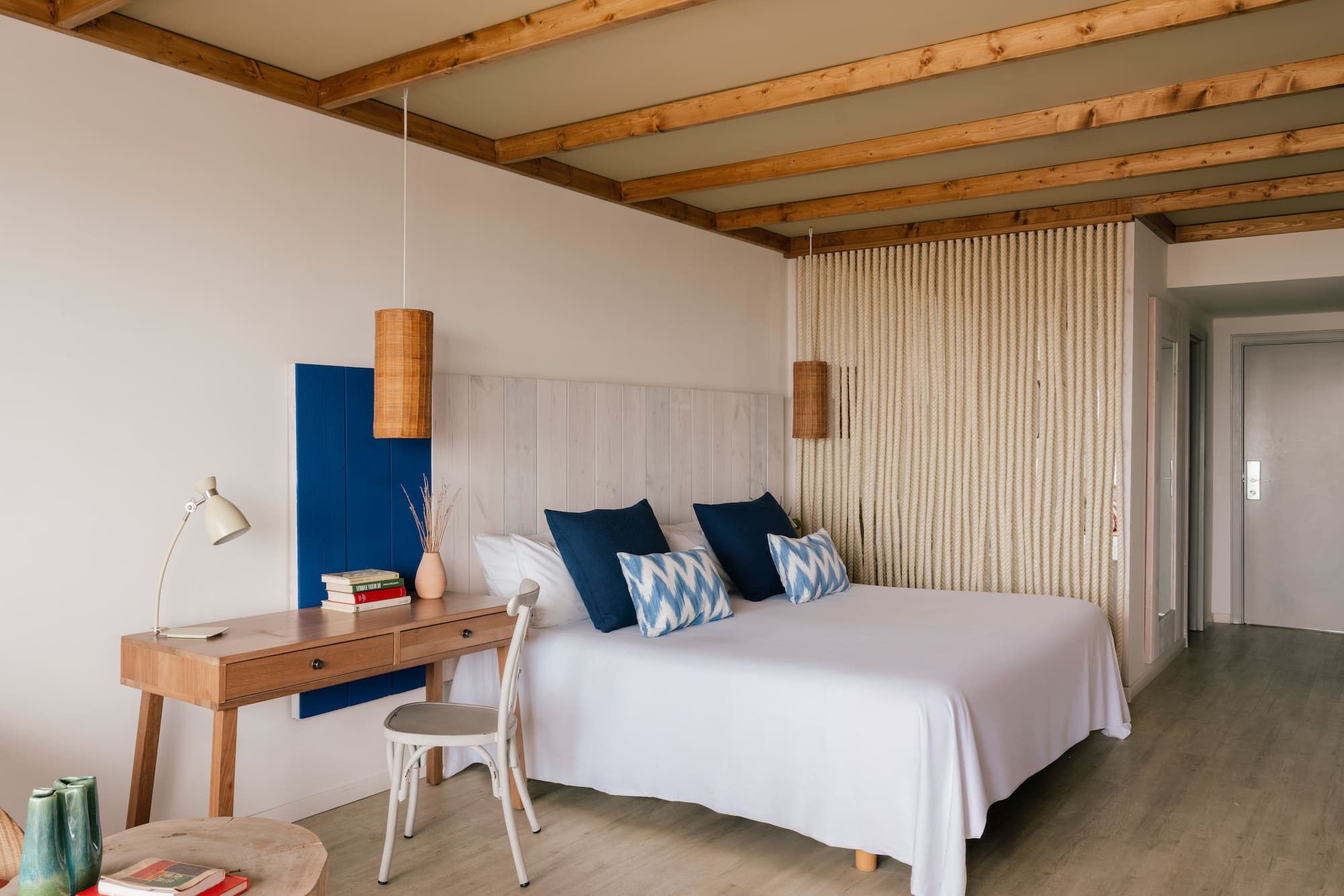 Dormitorio estilo mediterráneo con cama blanca, cojines azules, escritorio y vigas.