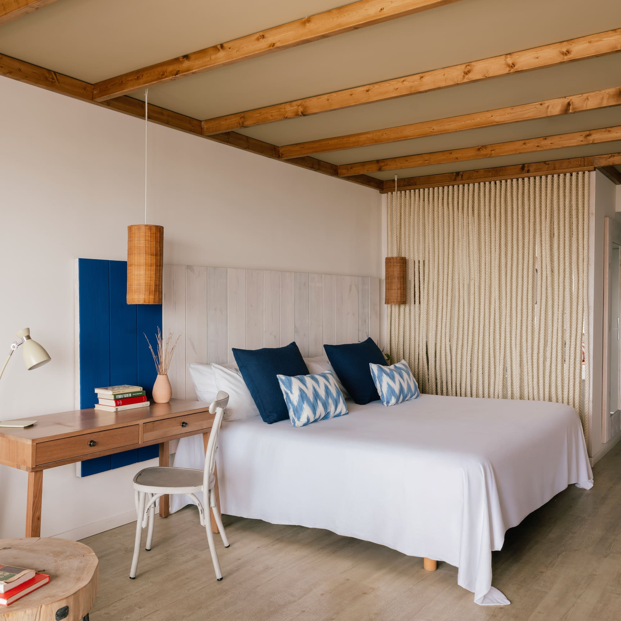 Dormitorio estilo mediterráneo con cama blanca, cojines azules, escritorio y vigas.