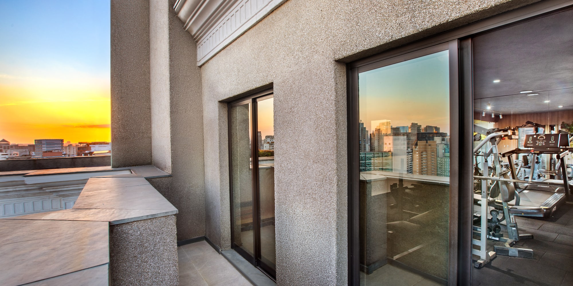 Apartment balcony with sunset city view, reflecting a modern gym with fitness equipment.