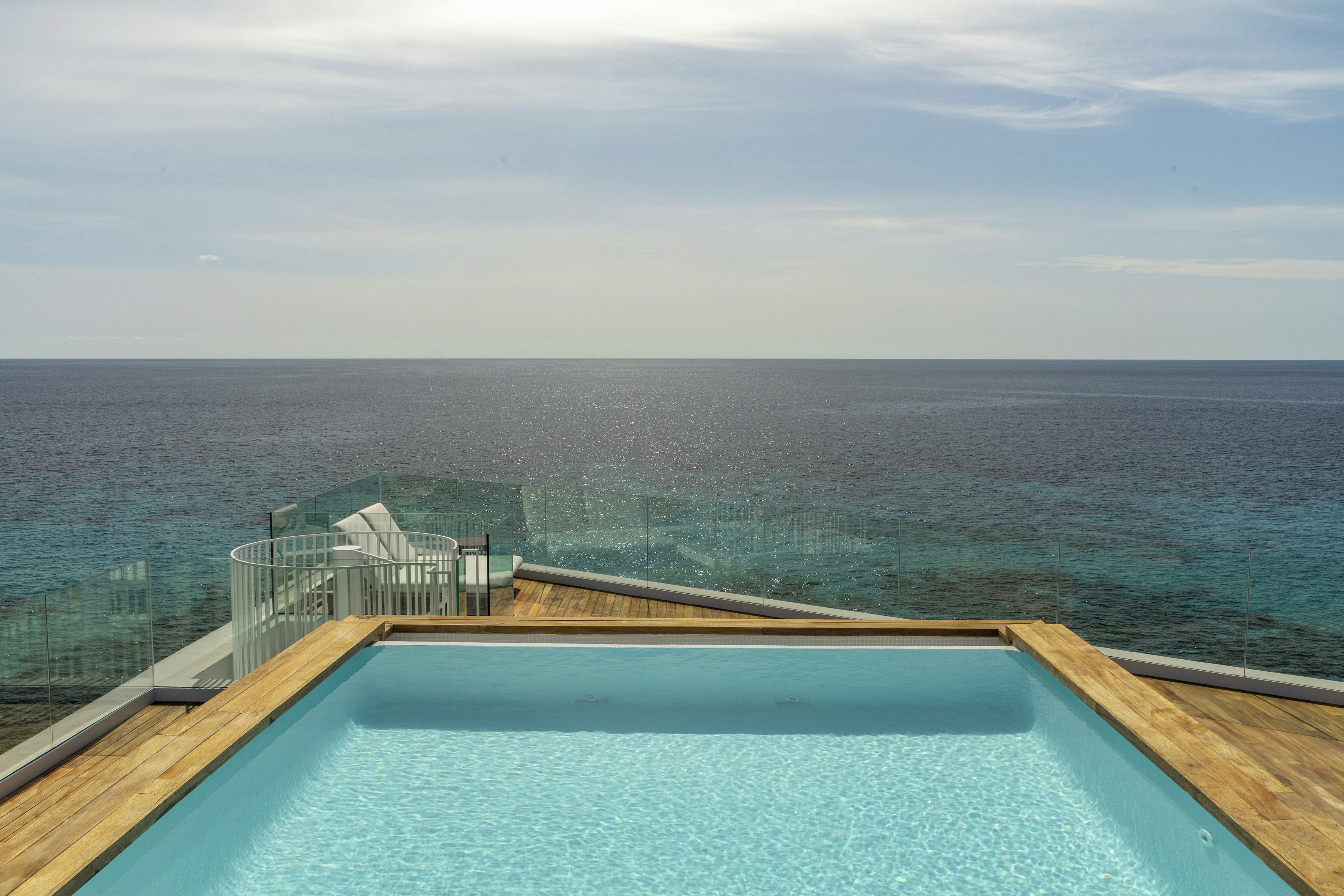 Infinity pool on wooden deck with glass railing overlooking ocean.