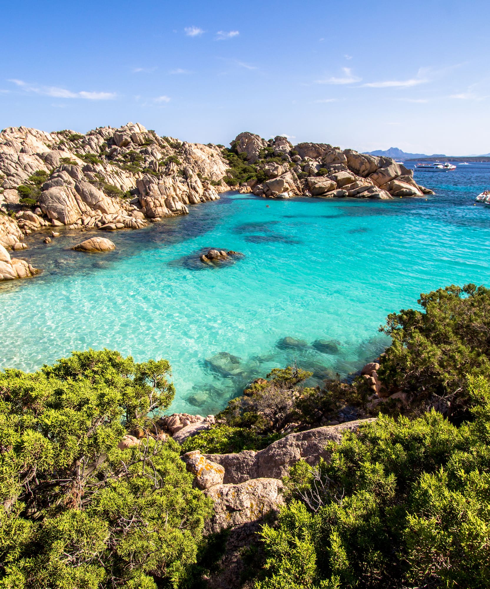 Clear turquoise bay, rocky shoreline, green shrubs, and distant boats.