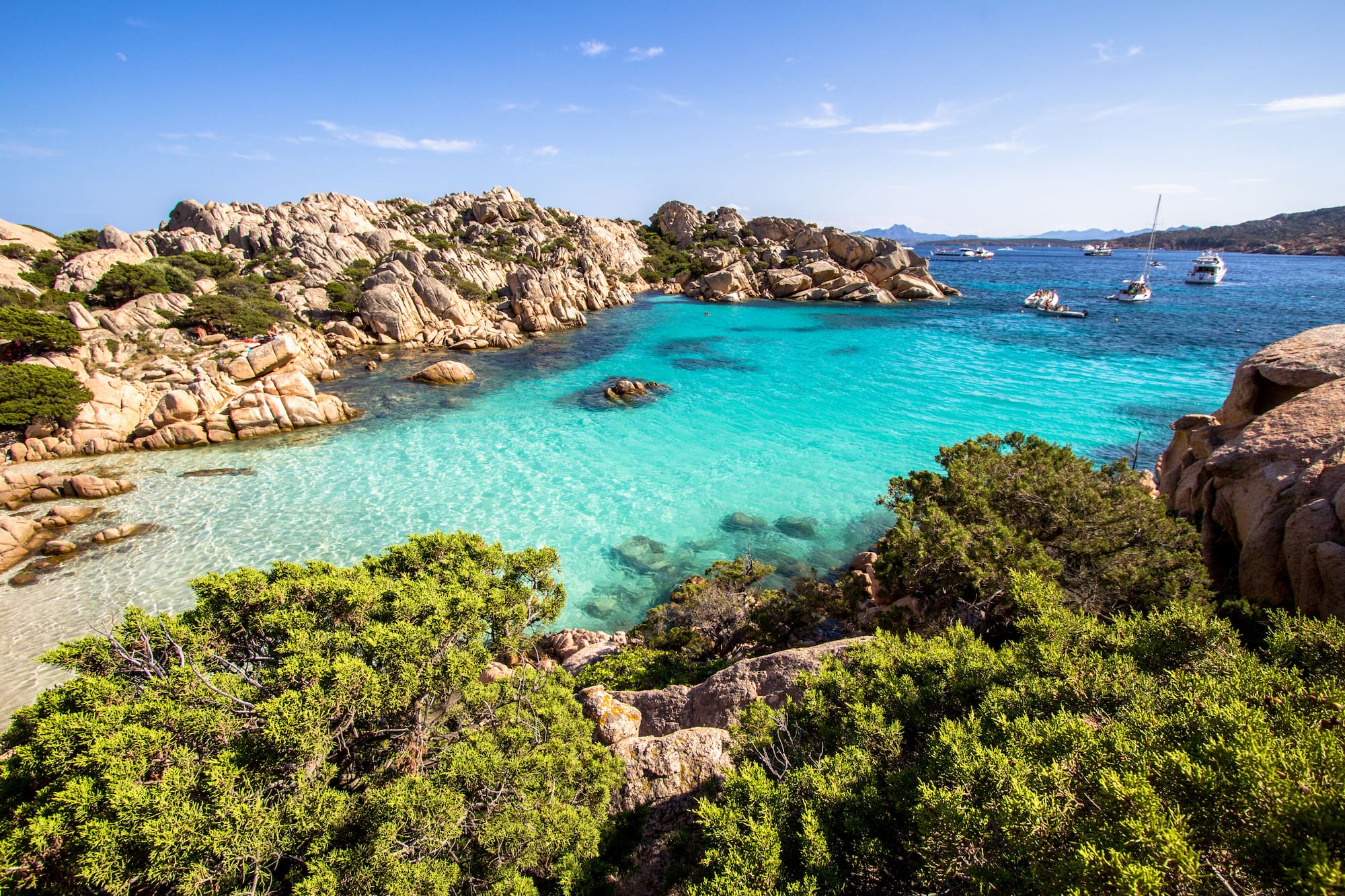 Clear turquoise bay, rocky shoreline, green shrubs, and distant boats.